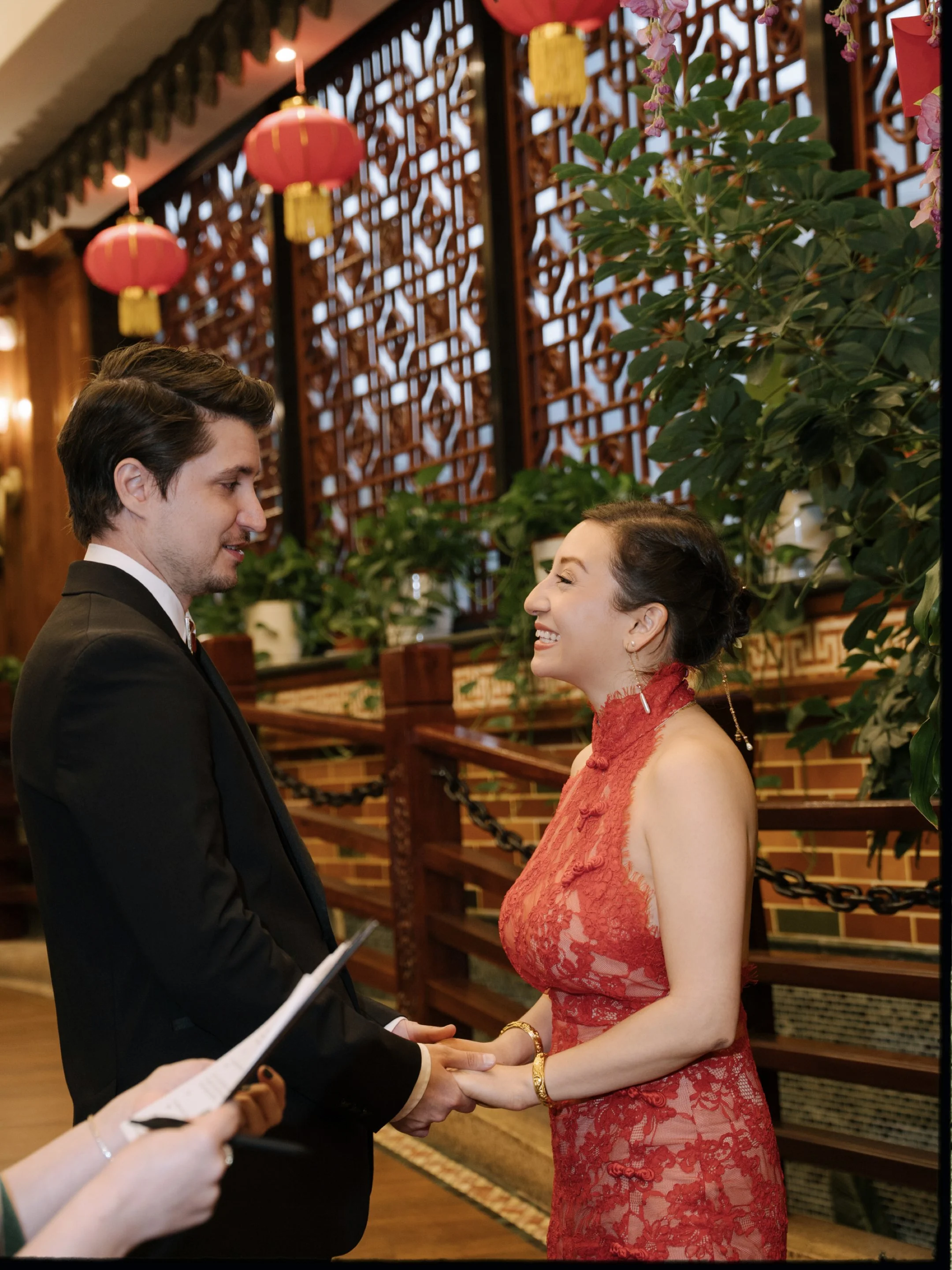 A man and woman face each other holding hands, in a setting decorated with red lanterns and traditional Chinese décor, likely during a wedding or formal ceremony.
