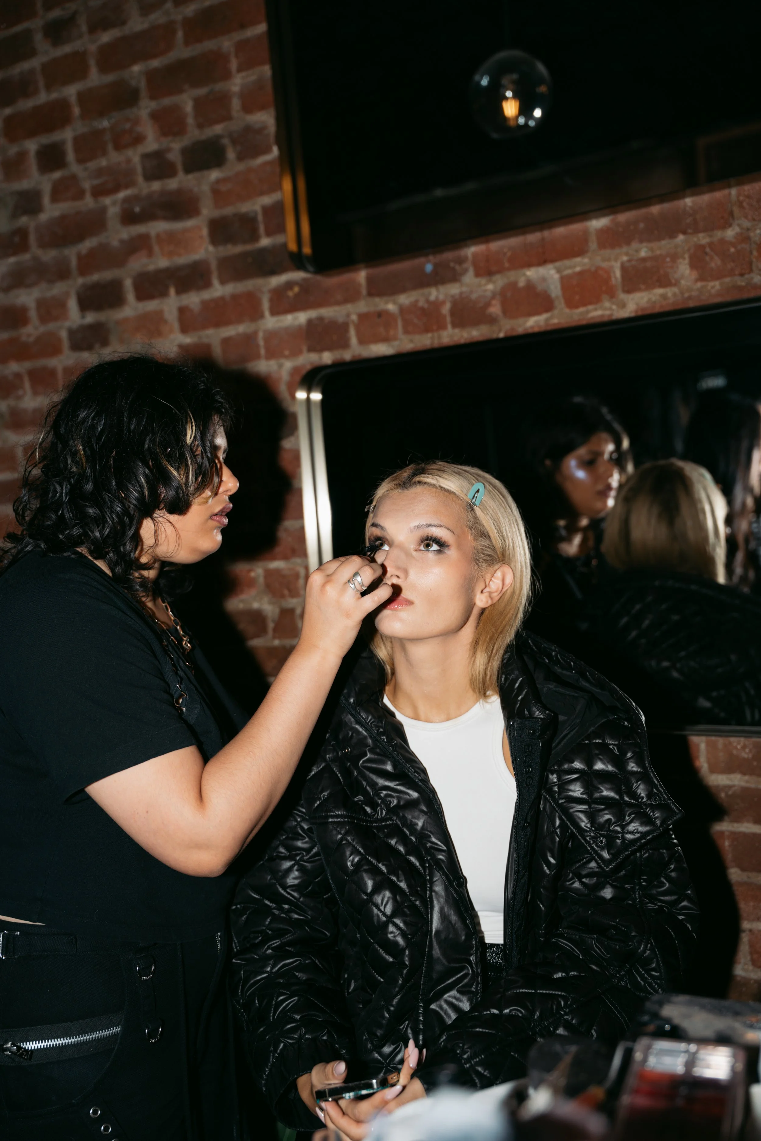 A makeup artist applies makeup to a blonde woman with blue eyes, wearing a black quilted jacket and white t-shirt, while another woman looks at her reflection in a mirror in a room with a brick wall.