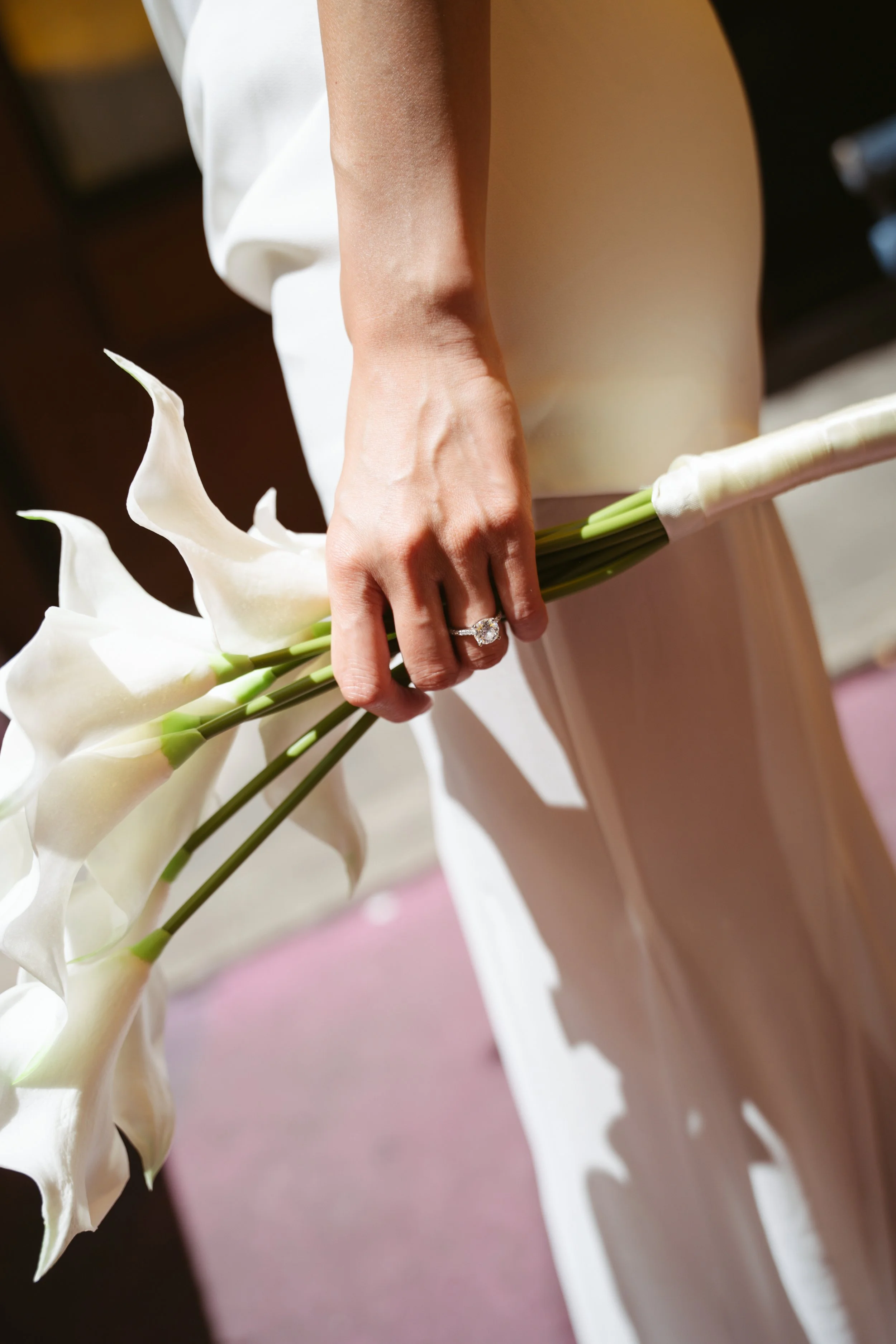 Close-up of a person's hand holding a bouquet of white calla lilies, with a diamond ring on their finger. The person is wearing a white dress.
