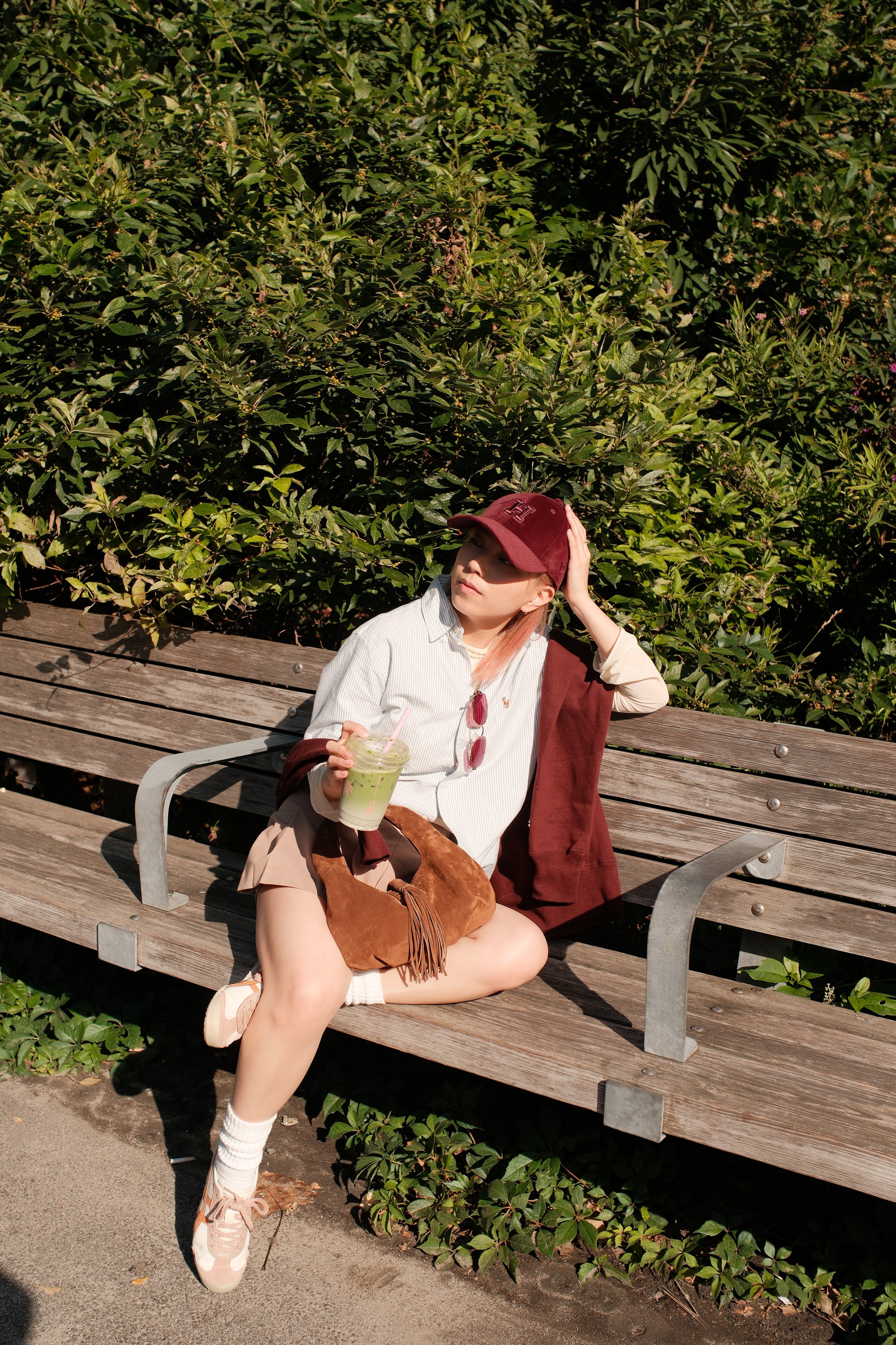 A young woman sitting on a wooden park bench with greenery in the background, wearing a burgundy cap, white shirt, beige shorts, and pink sneakers, holding a green smoothie in her right hand with sunglasses hanging from her shirt, and touching her ha