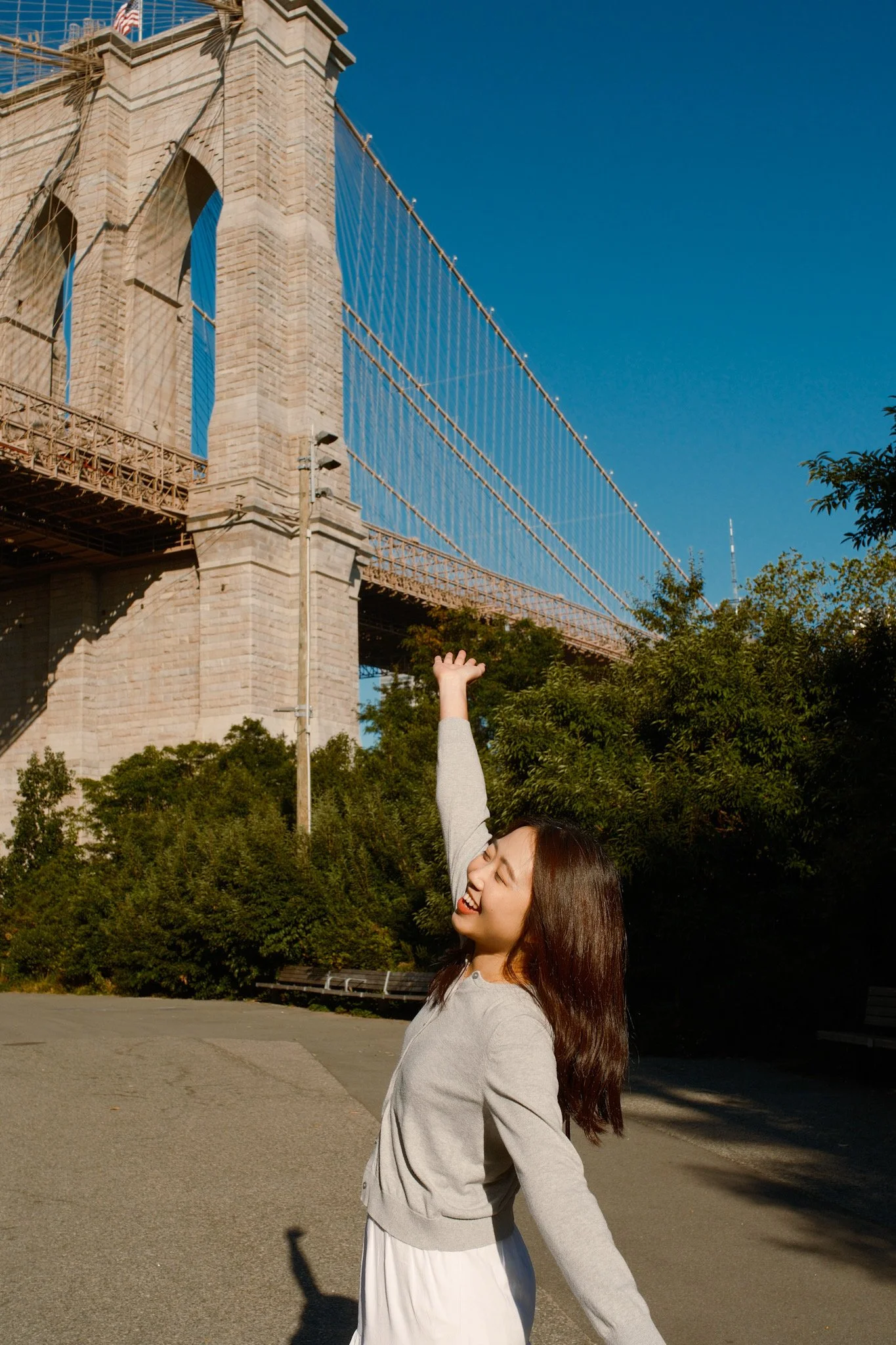 A woman smiling and raising her arm in front of the Brooklyn Bridge in New York City, with clear blue sky and trees in the background.