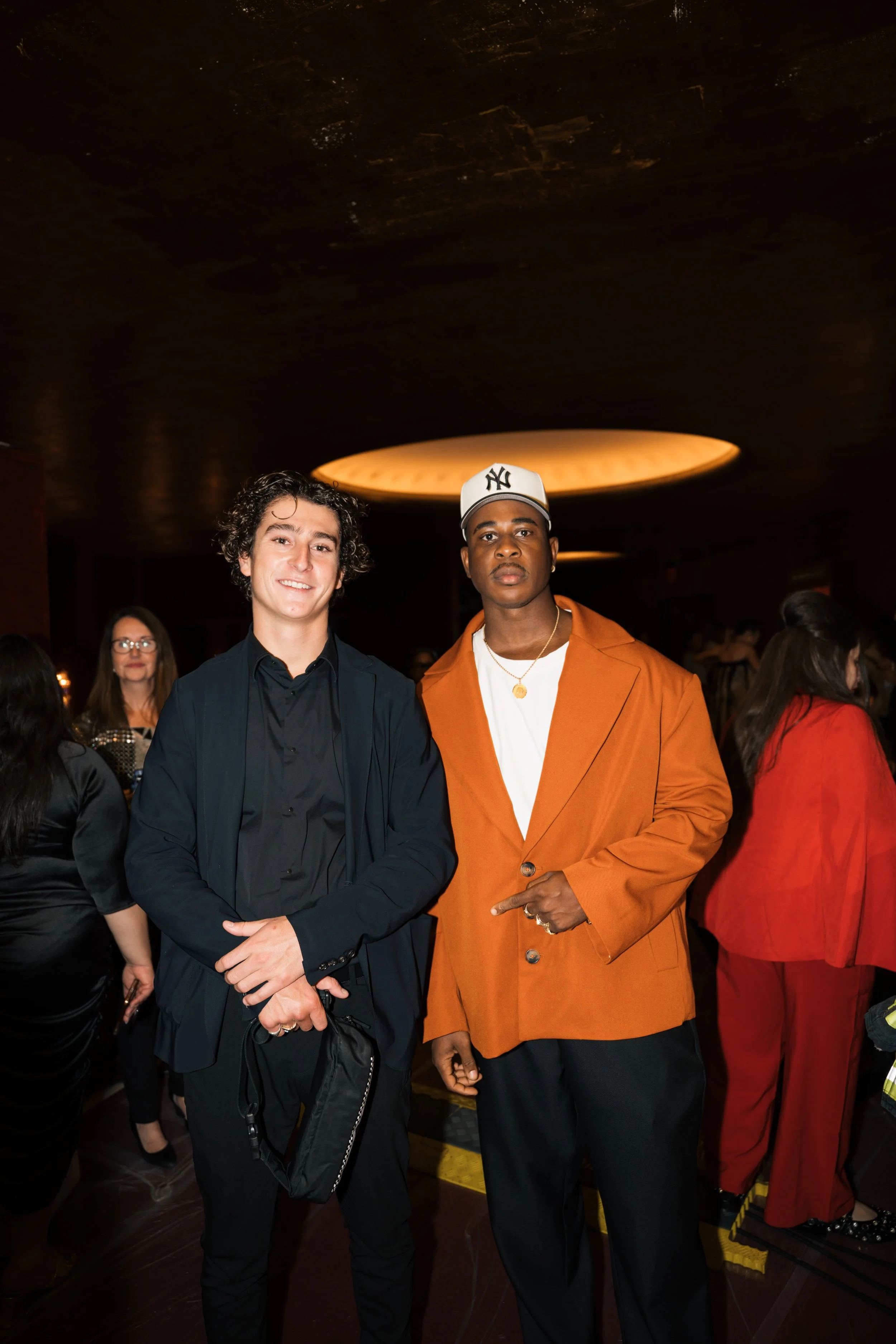 Two men posing together at a social event indoors. The man on the left has curly dark hair, a light complexion, and is wearing a black suit with a black shirt. The man on the right has a darker complexion and is wearing a white baseball cap with the 