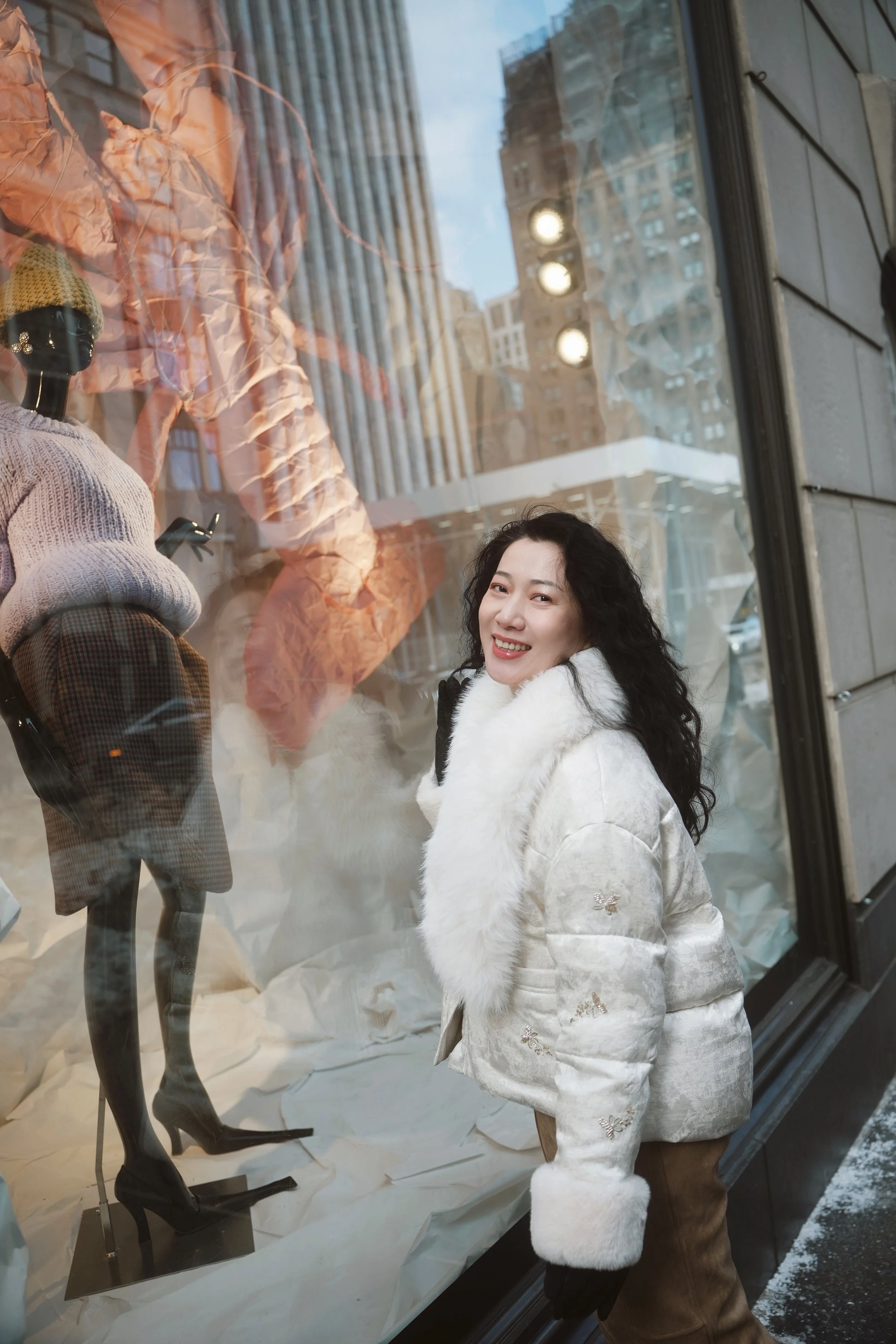 A woman with long dark curly hair smiling while standing outside a clothing store window display, which features a mannequin dressed in a gray sweater, plaid skirt, and a yellow beanie, with the cityscape reflected in the glass.