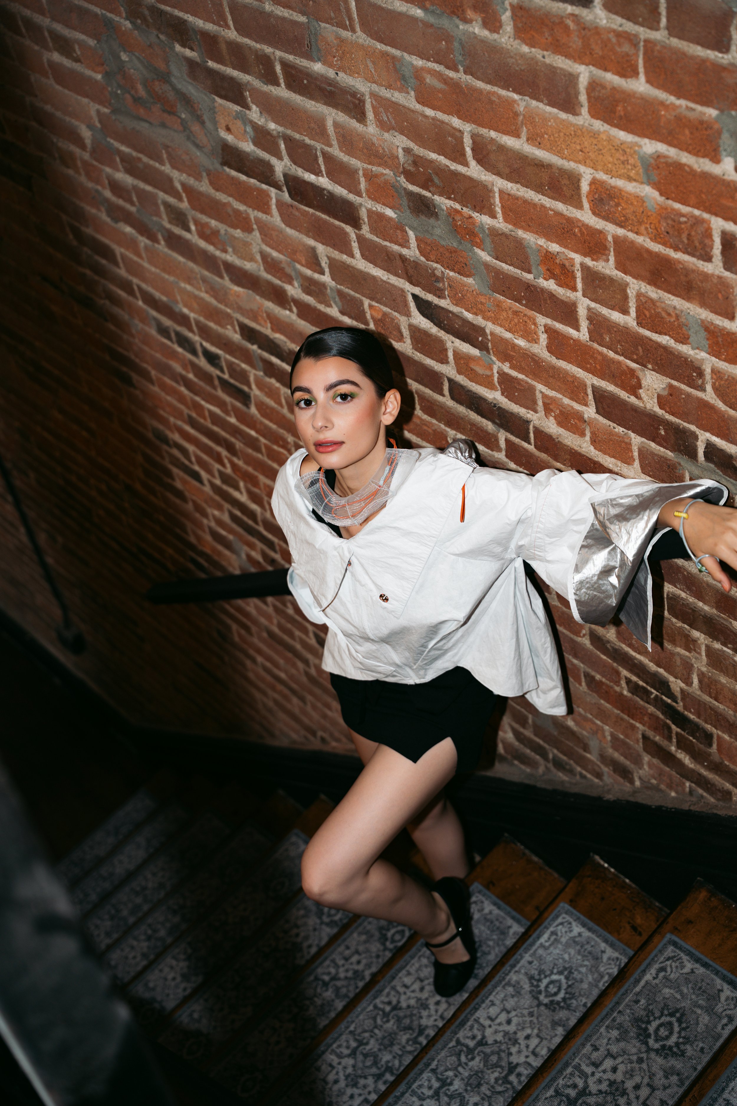 A woman with dark hair in a sleek style poses on a staircase with a brick wall background. She wears a white jacket, black shorts, and black shoes, with one arm extended along the staircase railing and the other resting on the wall.