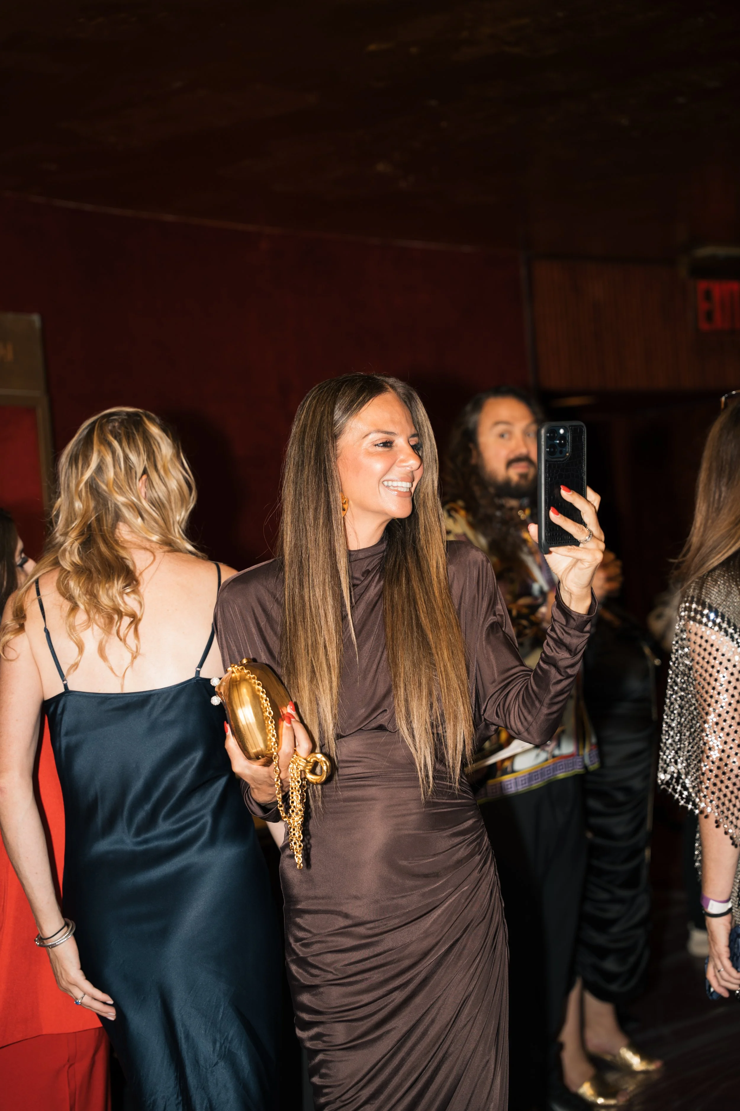 A group of women dressed in elegant evening wear at a social event. The woman in the center is smiling and taking a selfie with her phone, holding a gold clutch purse, with long brown hair and wearing a metallic brown dress. Other women around her ar