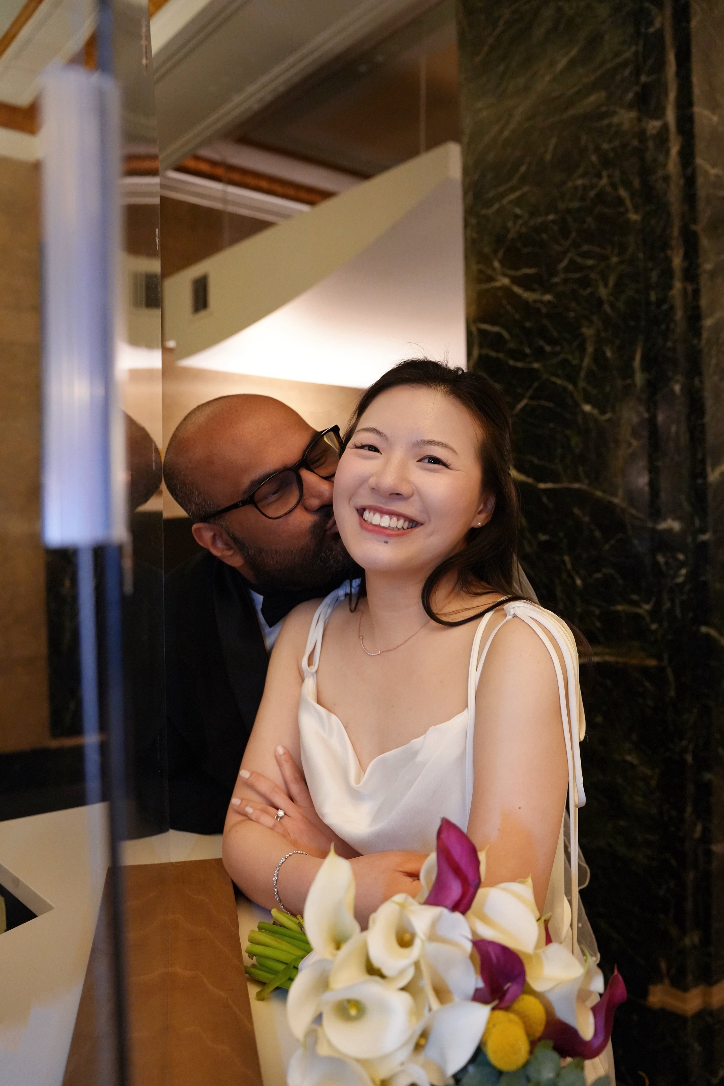 A man and woman sharing a joyful moment near a flower arrangement at a celebration. The woman is smiling at the camera, and the man is giving her a gentle kiss on the cheek.
