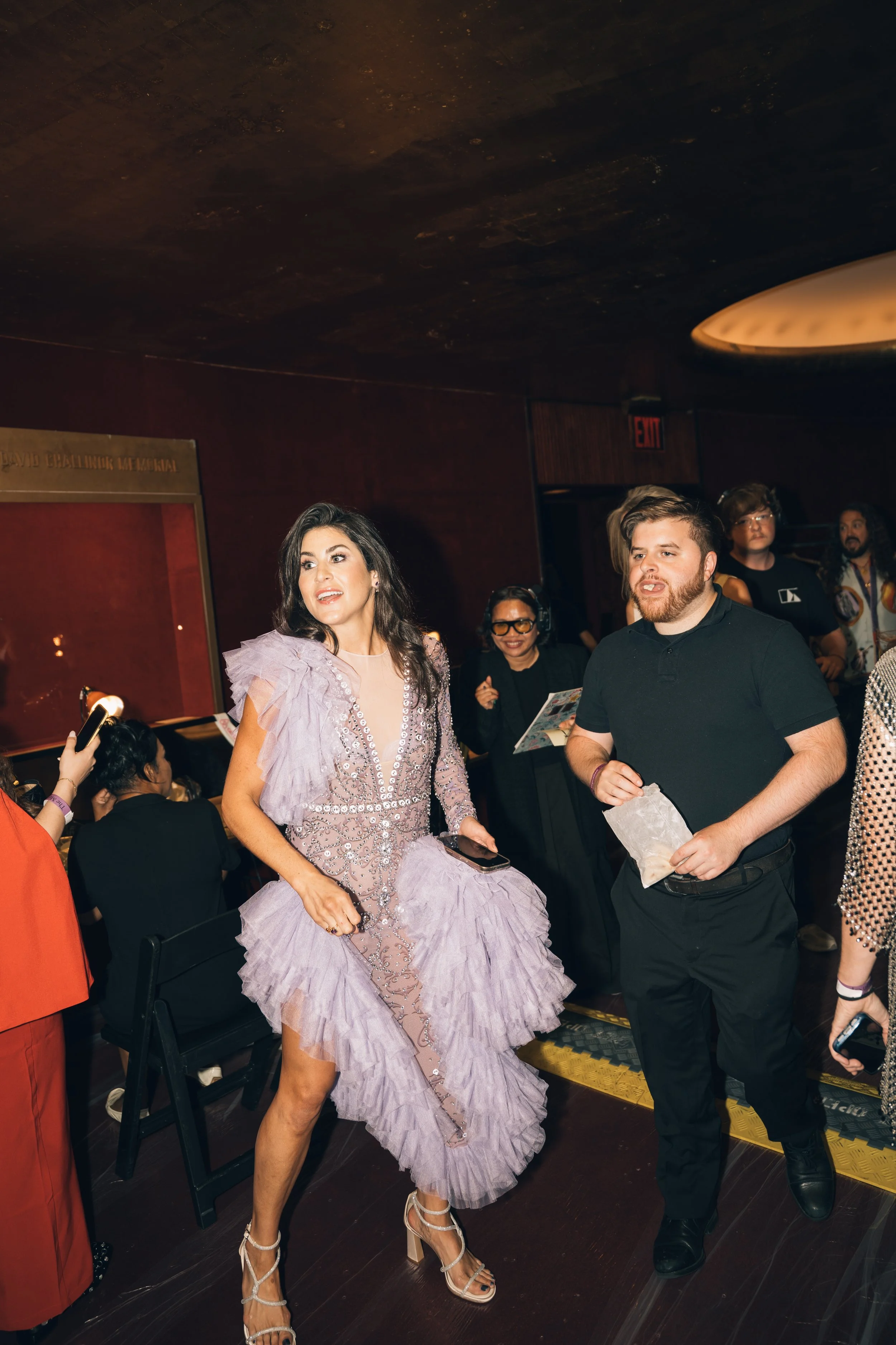A woman dressed in an elegant lavender gown with intricate beading and ruffles, wearing high heels, is standing in a dimly lit room with dark walls. There are several other people in the background, some with headsets, and a man in a black shirt hold