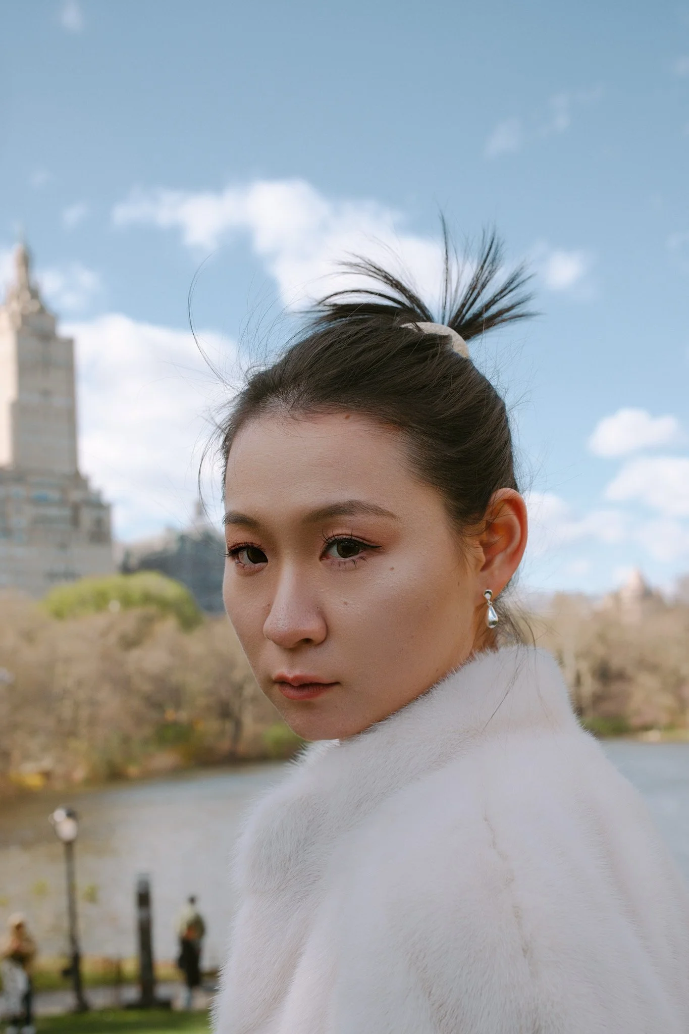 Close-up of a woman with dark hair tied up in a high ponytail, wearing a white turtleneck sweater, with a cityscape and river in the background under a partly cloudy sky.