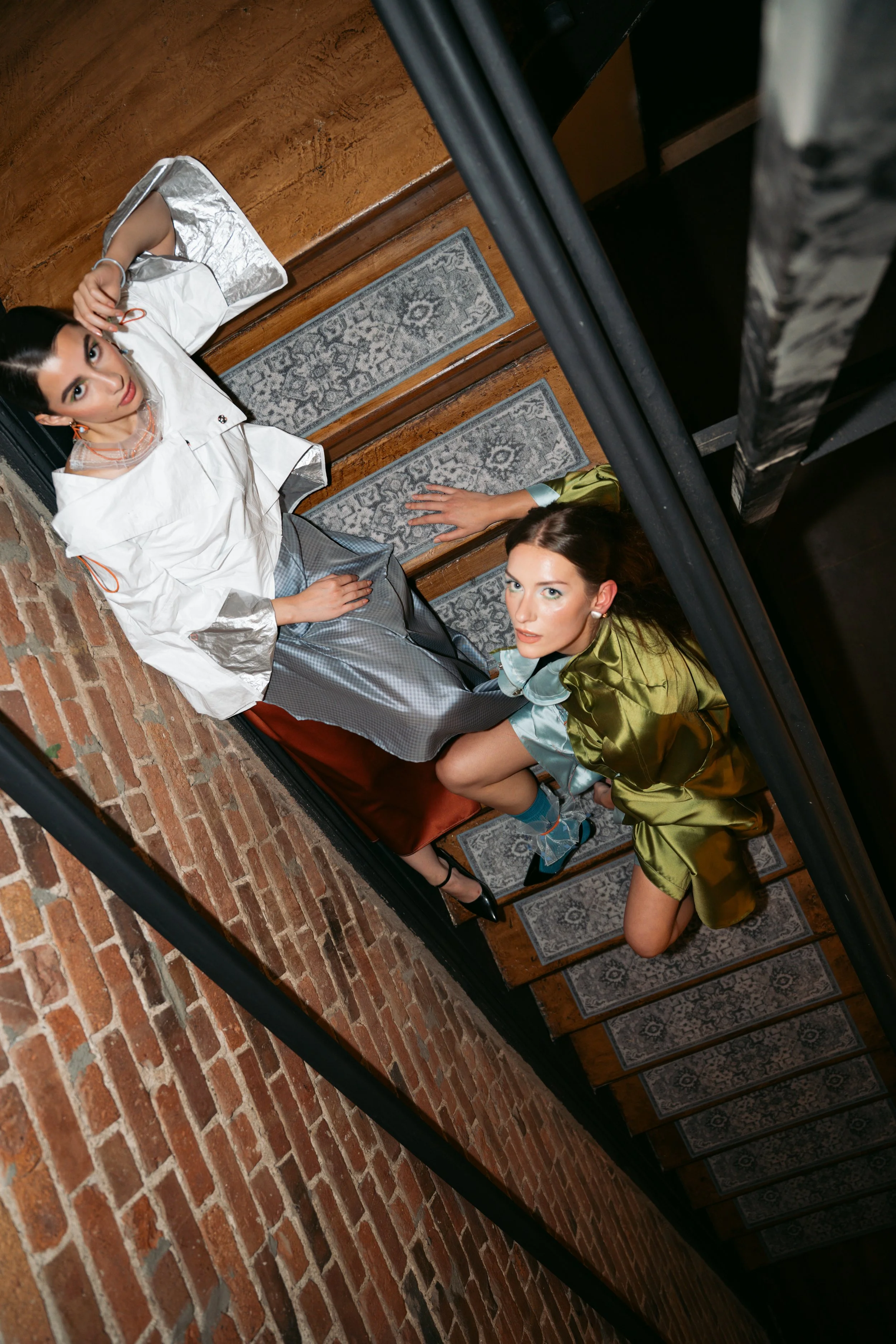 Two women with dark hair and fair skin, one in a white shirt and the other in a satin green shirt, looking up at a camera from the stairs in a rustic scene with brick walls and patterned rugs.