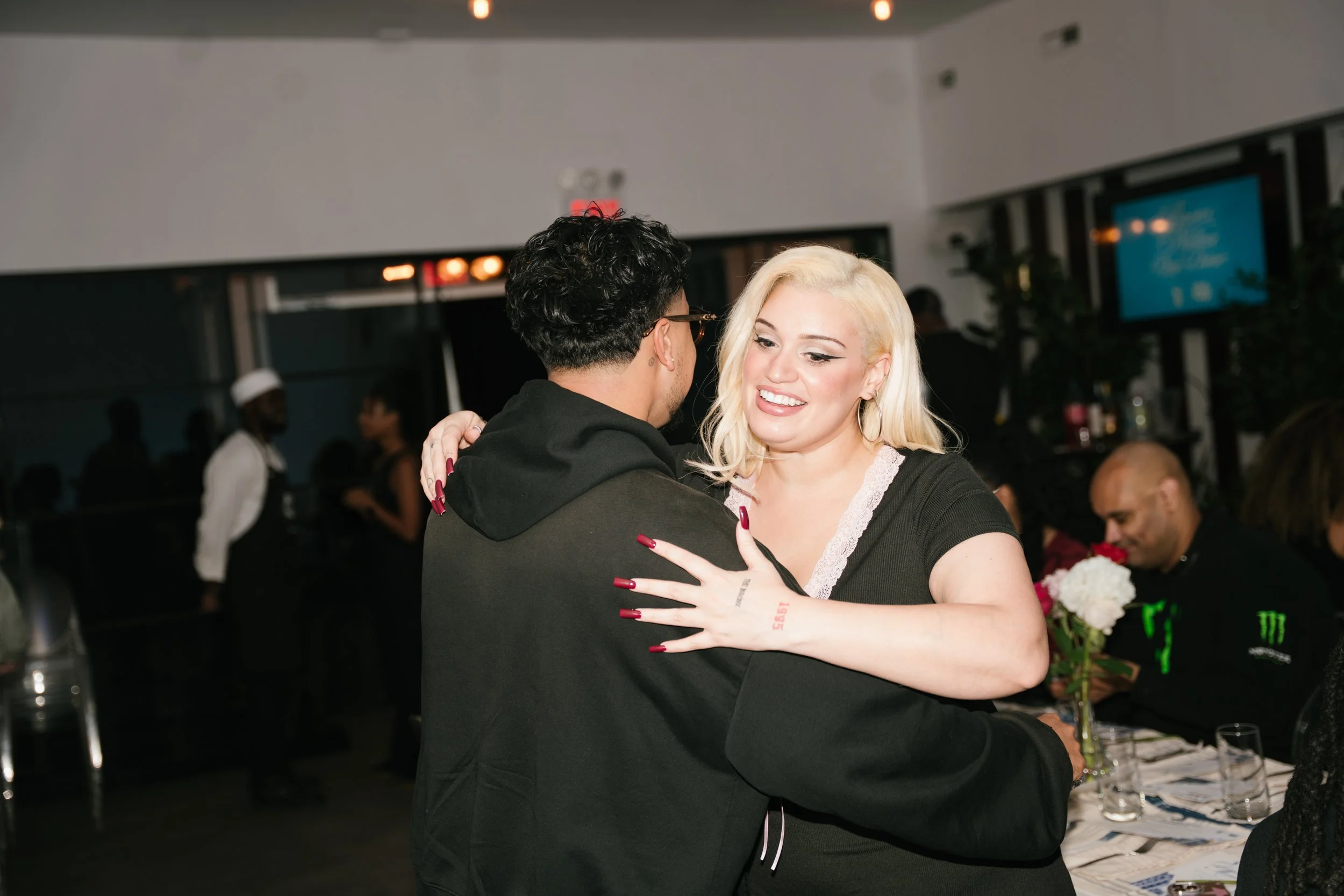 A man and woman dance closely together at a celebration or party, smiling and holding each other in an embrace. In the background, there are other people, some seated at a table with flowers, and a bartender serving drinks. The setting appears to be 