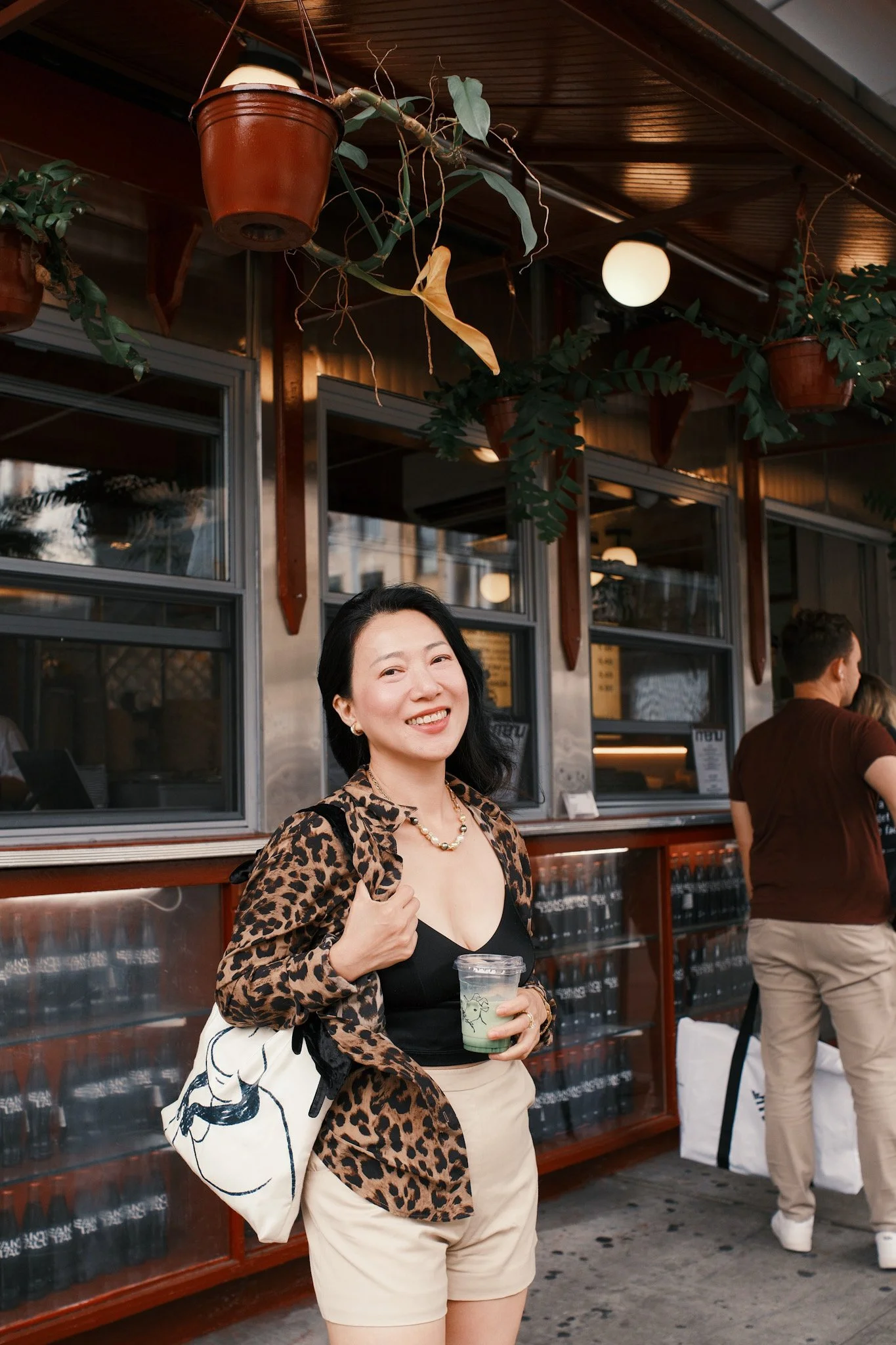 A woman with dark hair and earrings smiling while holding a plastic cup with a green drink, wearing a leopard print jacket over a black top and beige shorts. She is standing outdoors in front of a wooden and glass building, with hanging plants and li