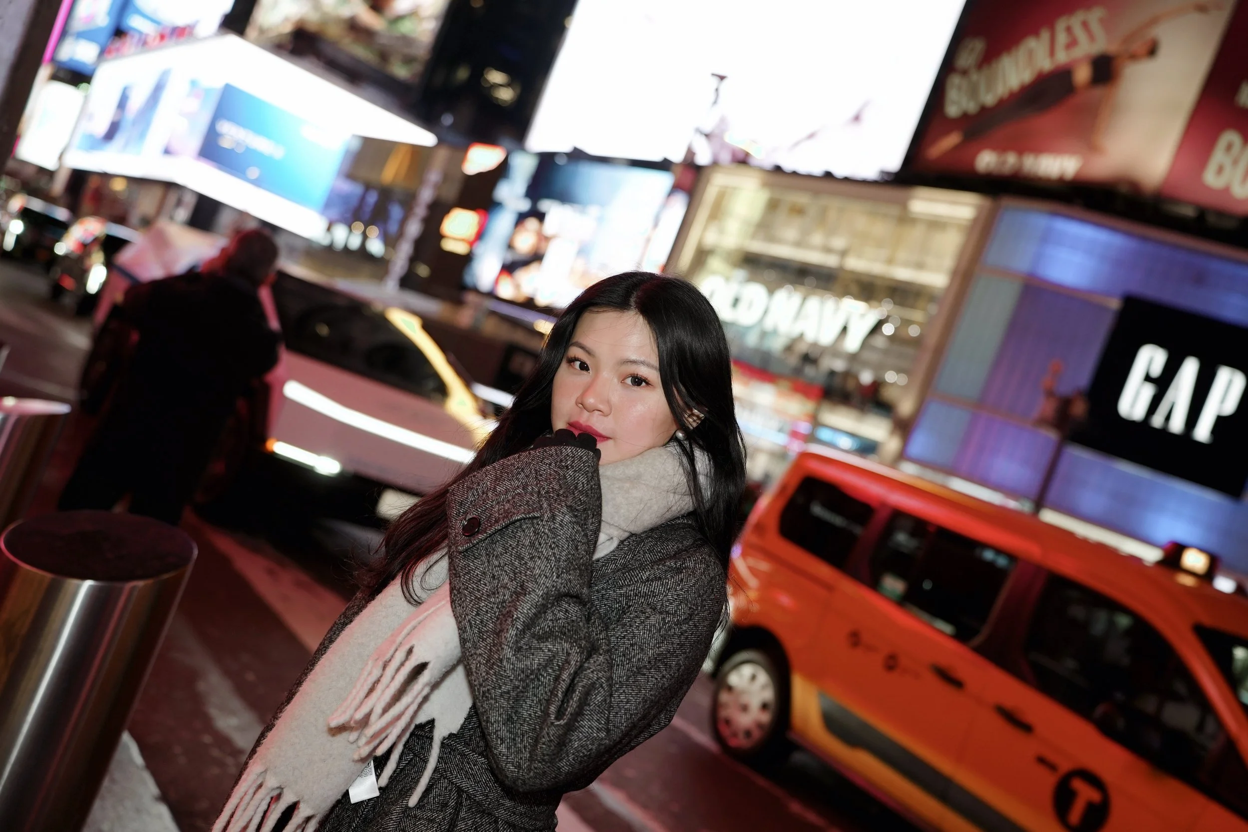 A young woman with long dark hair wearing a wool coat and a beige and pink scarf standing at a city street at night, with bright billboards and a yellow cab in the background.