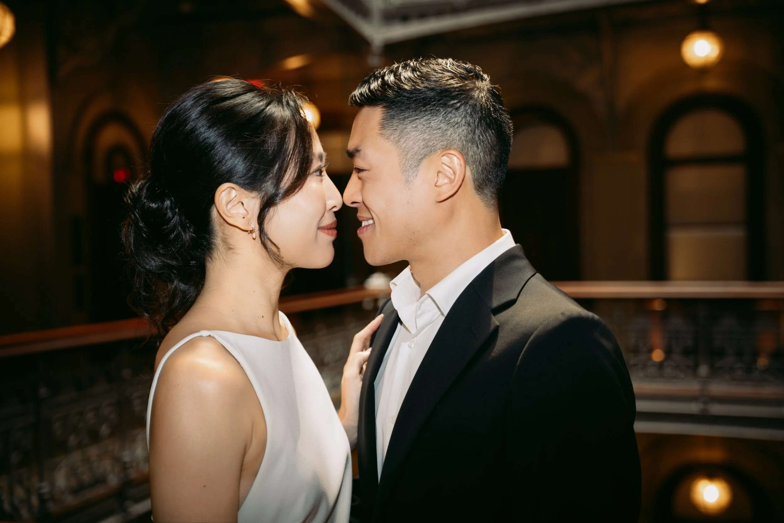A couple, dressed formally, face each other closely in an intimate moment indoors with warm lighting.