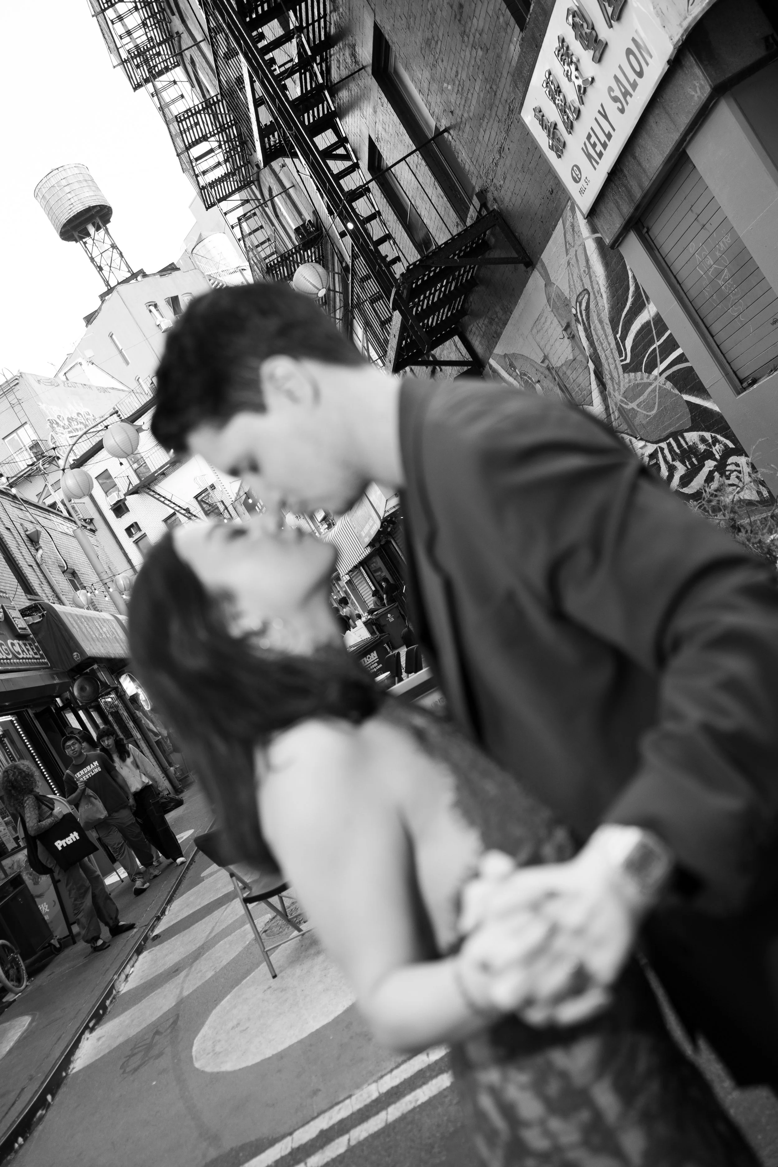 A black and white photo of two young people sharing a kiss outdoors on a city street, with various storefronts, pedestrians, and a water tower in the background.