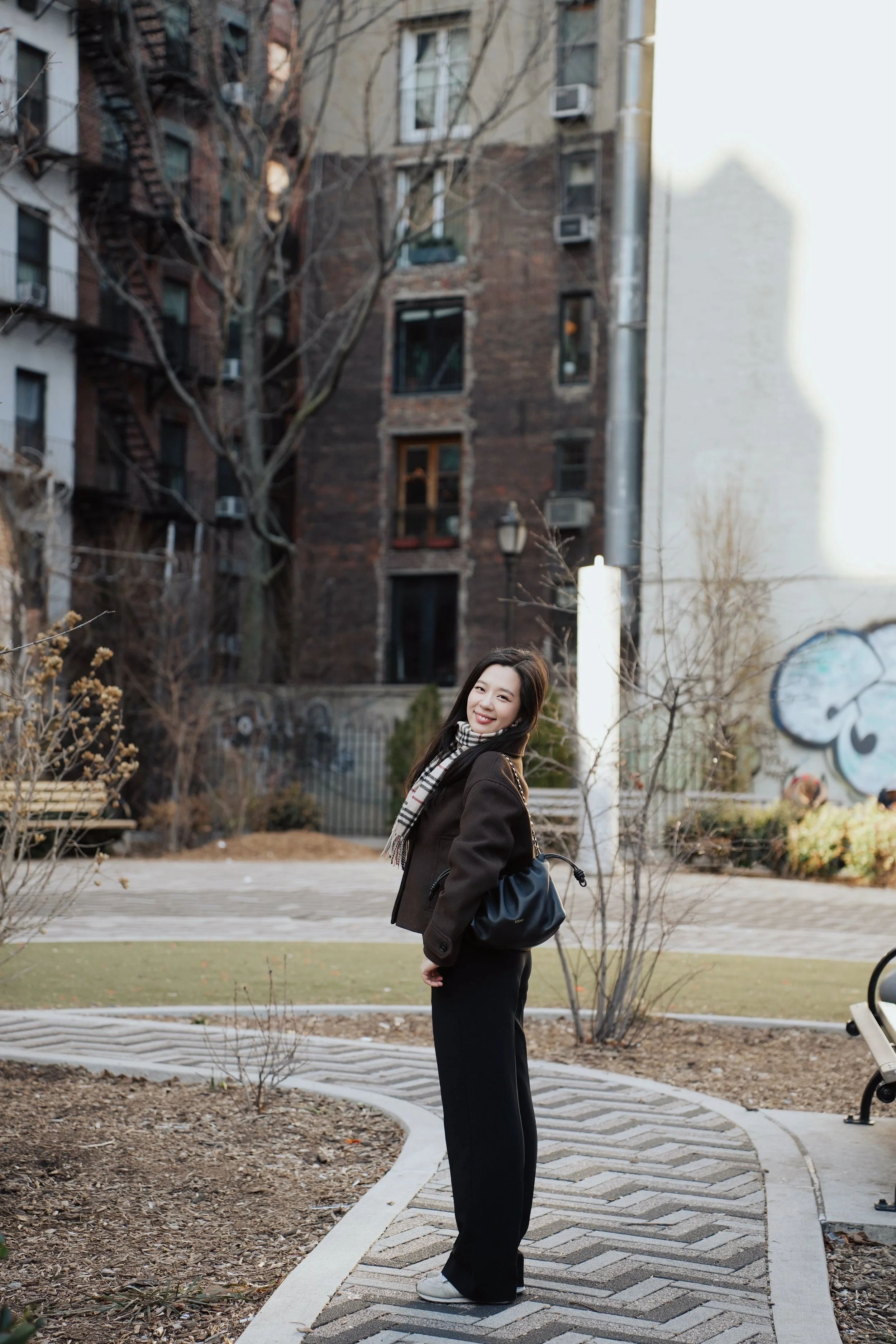A young woman smiling and looking at the camera, standing outdoors on a sidewalk with leafless trees and apartment buildings in the background.