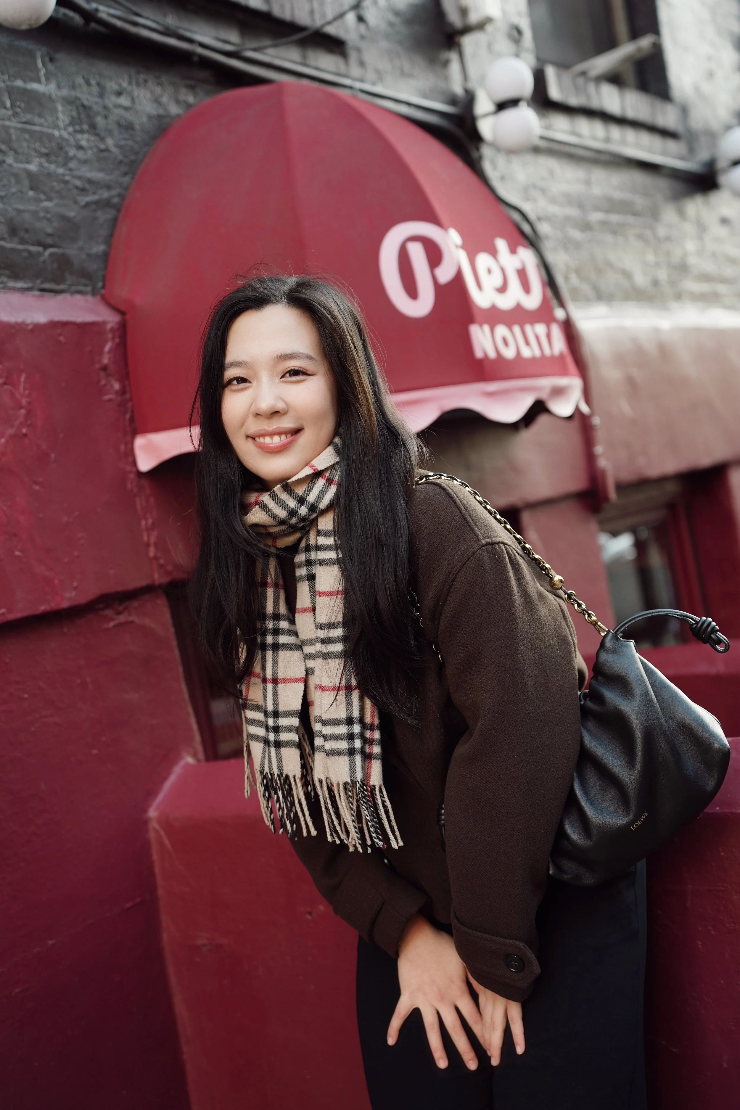 Close-up of a young woman with long dark hair wearing a plaid scarf, dark coat, and black pants, standing in front of a red wall with a sign that reads 'Petit Nolita'.