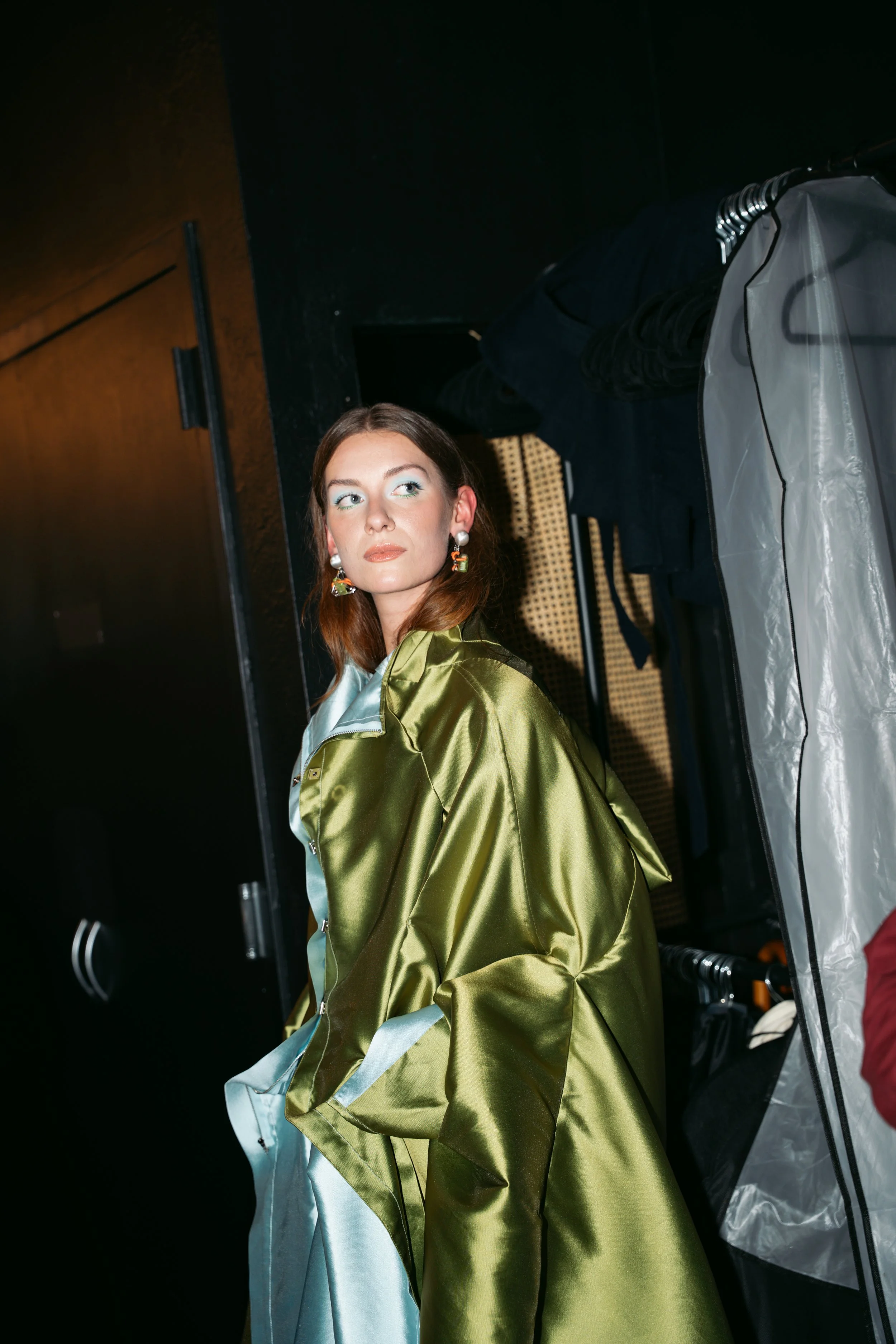 A woman with long brown hair, wearing colorful earrings and a shiny green jacket, standing behind a clothing rack.