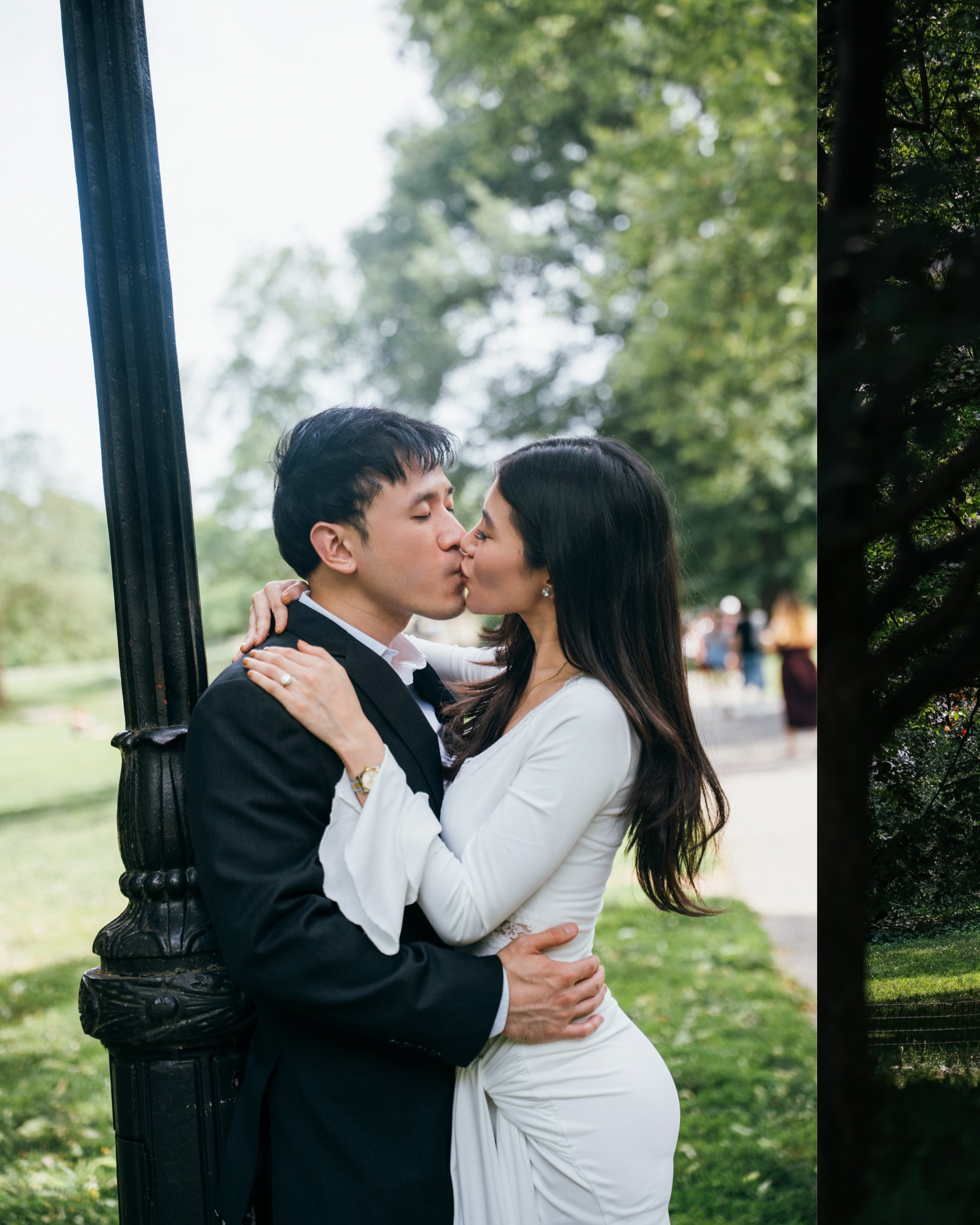 A couple is sharing a kiss outdoors, with the man in a suit and the woman in a white dress, standing near a black lamppost with lush green trees in the background.