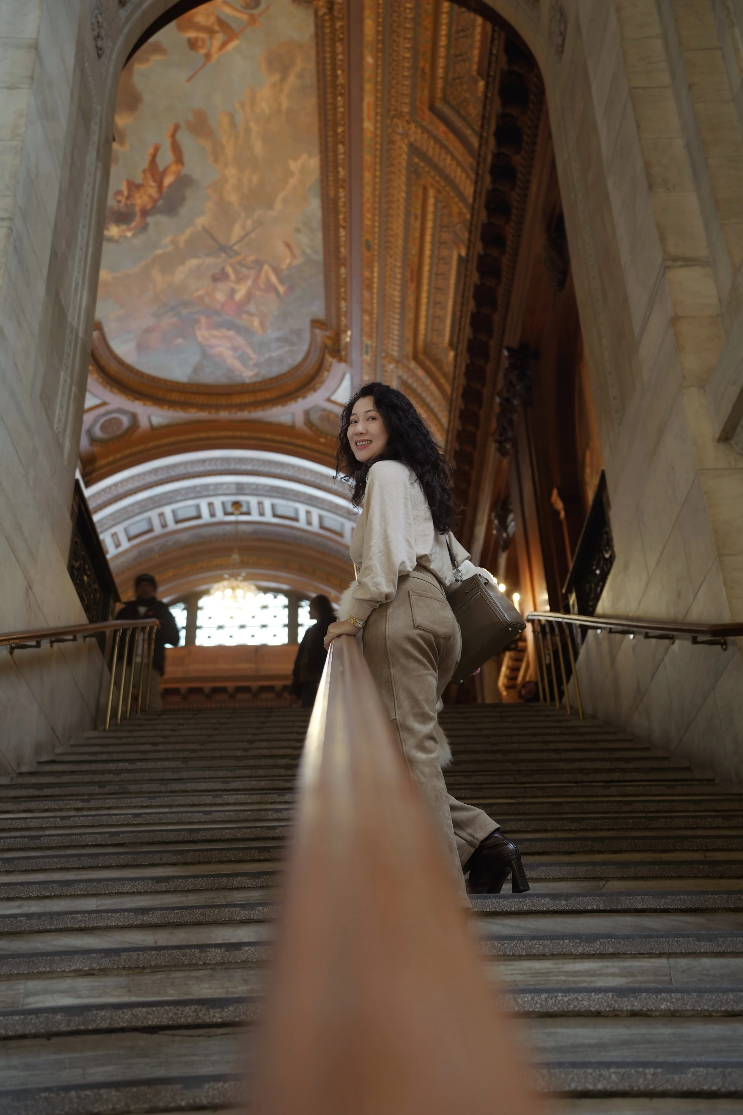 A woman with curly dark hair smiling and looking back while standing on the stairs inside a grand, ornate building with a painted ceiling and golden decorative elements.