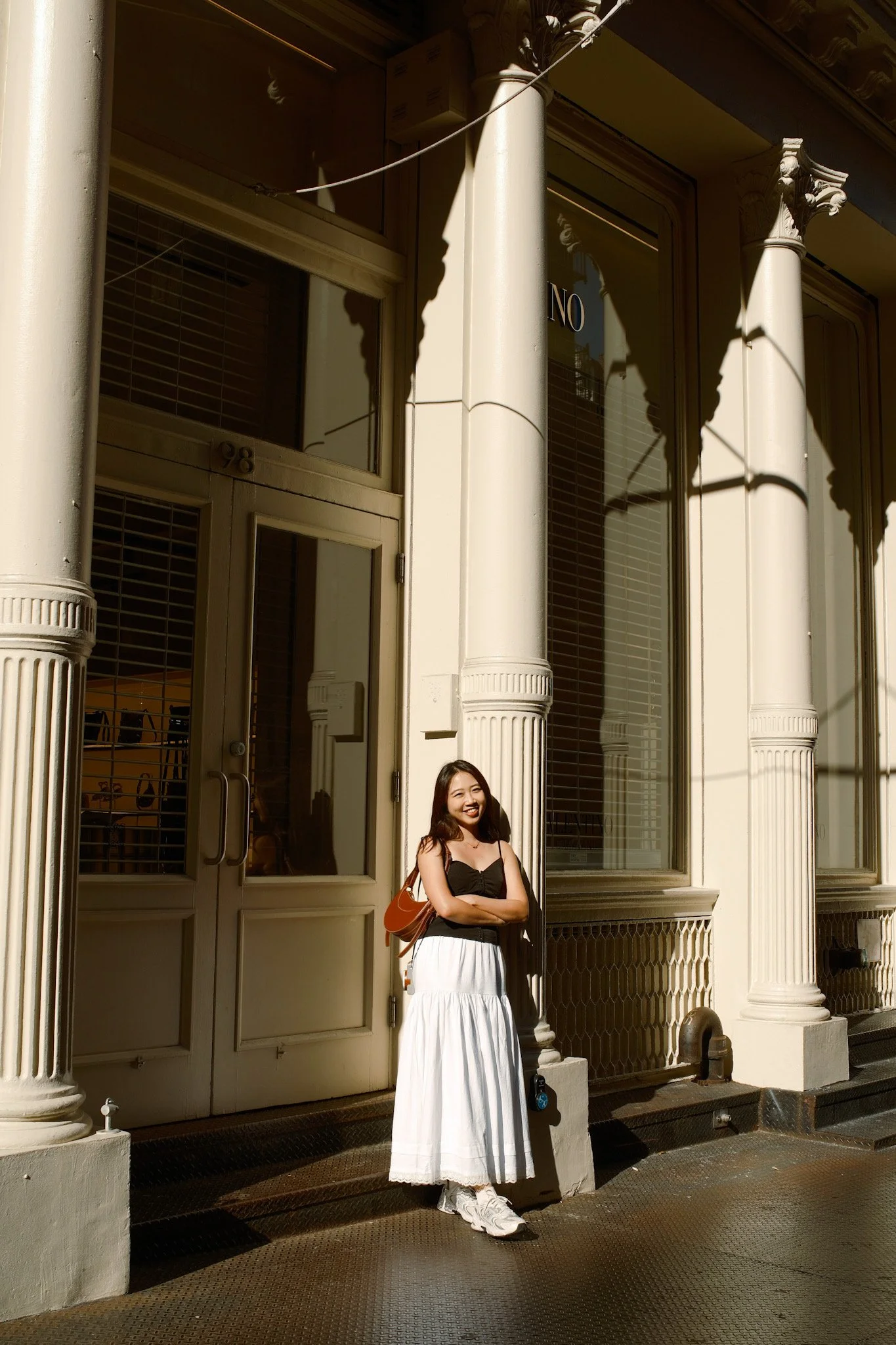 A young woman stands outside a building with tall columns and large windows, smiling and wearing a black top and a white skirt. She has a brown backpack and white sneakers, with sunlight casting shadows on the building facade.