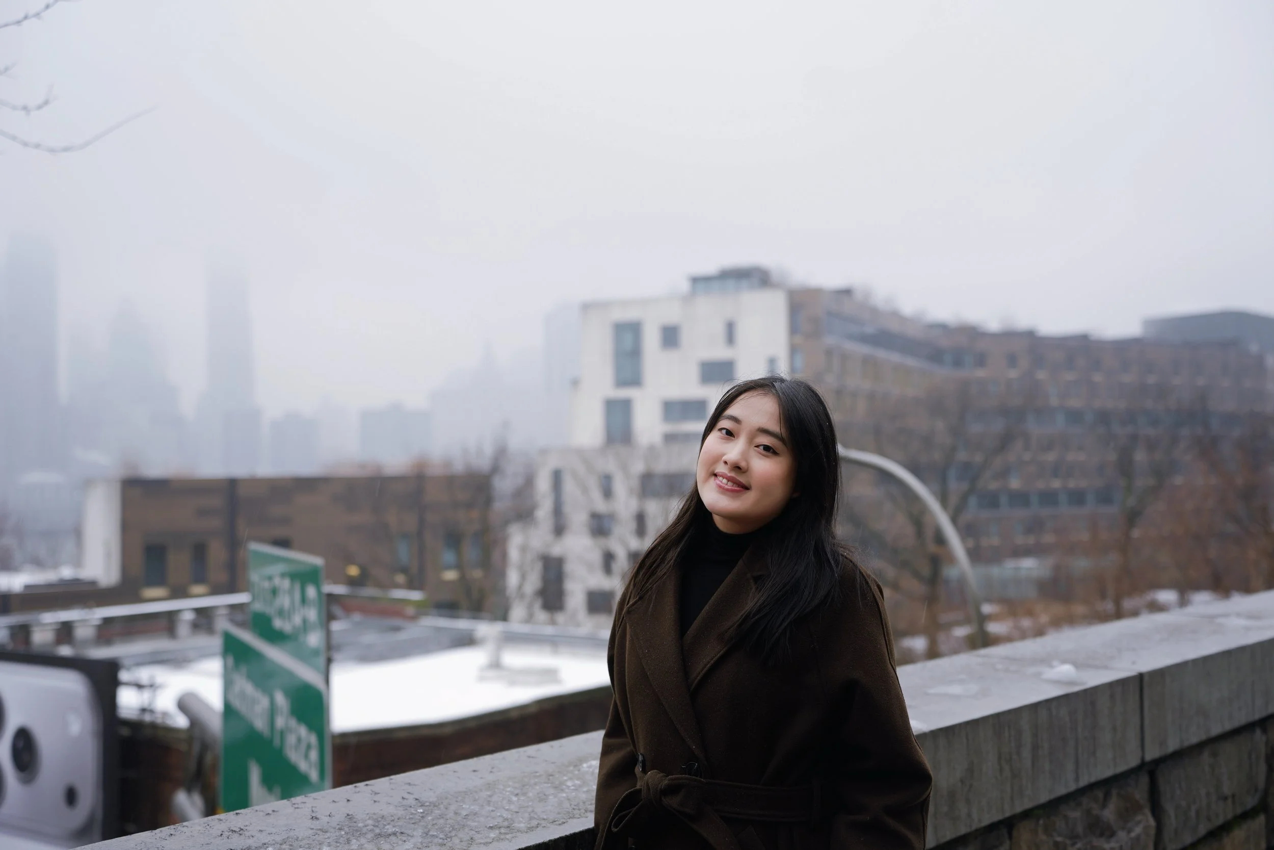 A young woman with long black hair standing outdoors on a cloudy day in an urban area with modern buildings in the background.