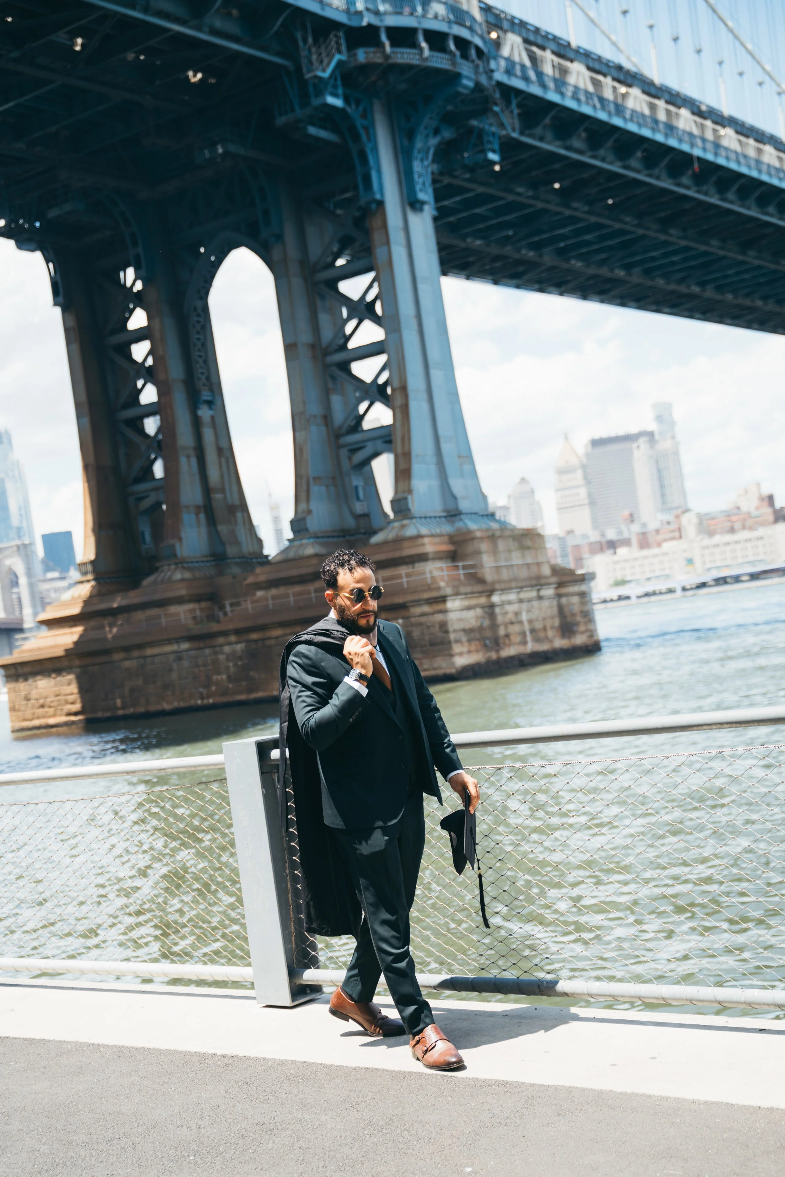 A man in a business suit walking along a riverside path under a bridge in an urban area, holding a briefcase and wearing sunglasses.