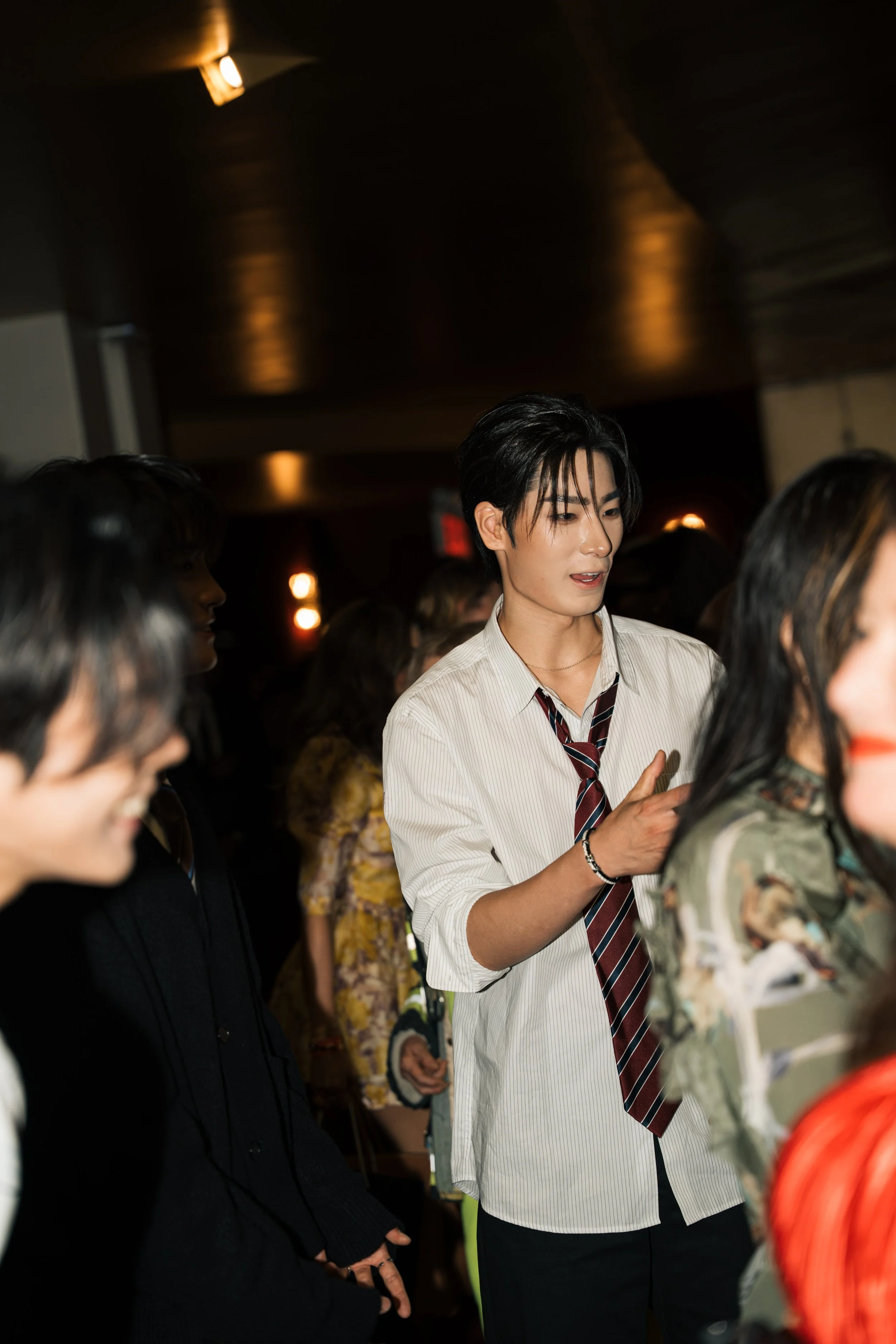 A group of people at an indoor event, with a focus on a young man in a white dress shirt and striped tie, engaging in conversation. The atmosphere is dimly lit with warm lighting.