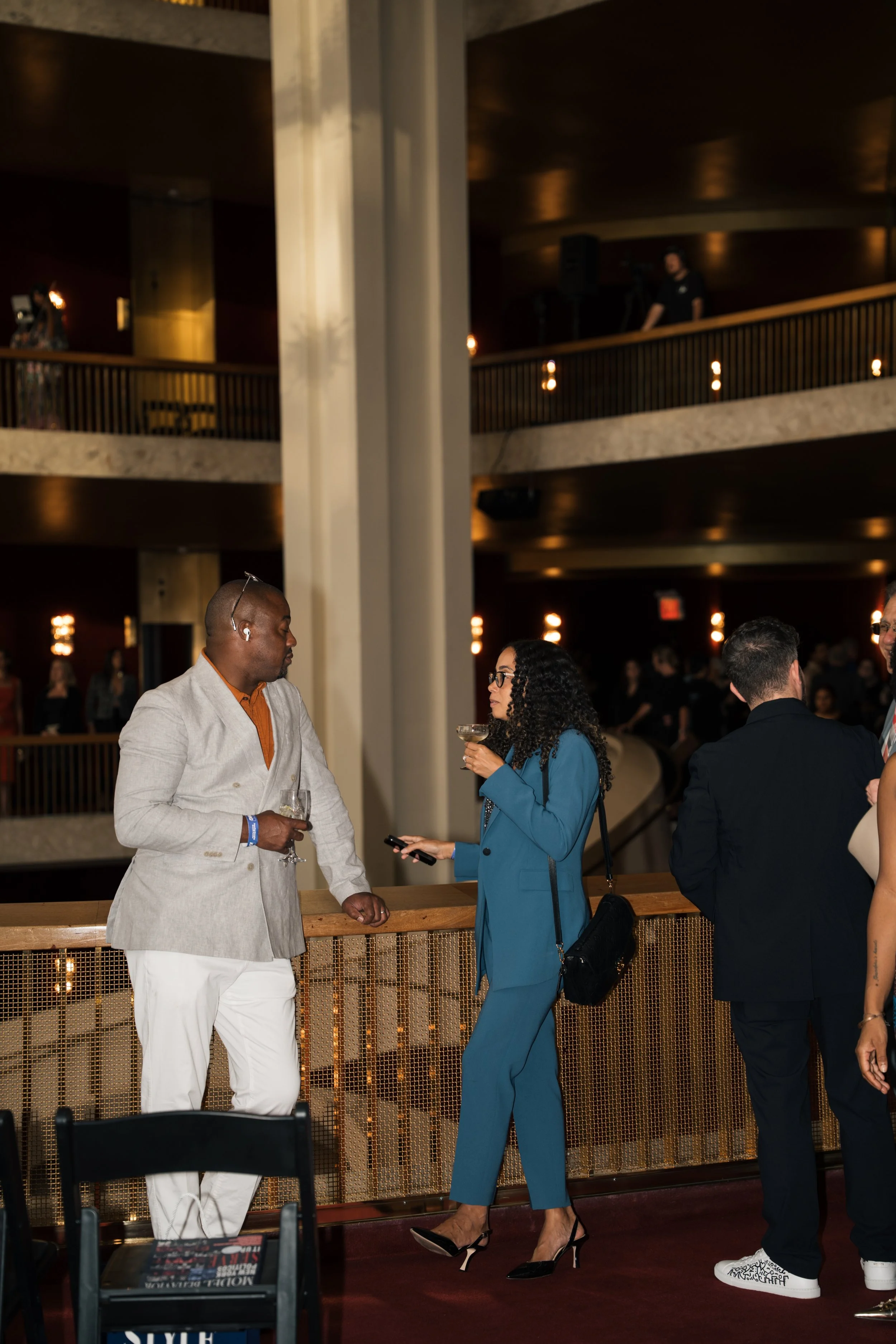 People are standing and talking at an indoor event or venue, with some holding drinks. The setting has warm lighting, a balcony or upper level, and modern decor.