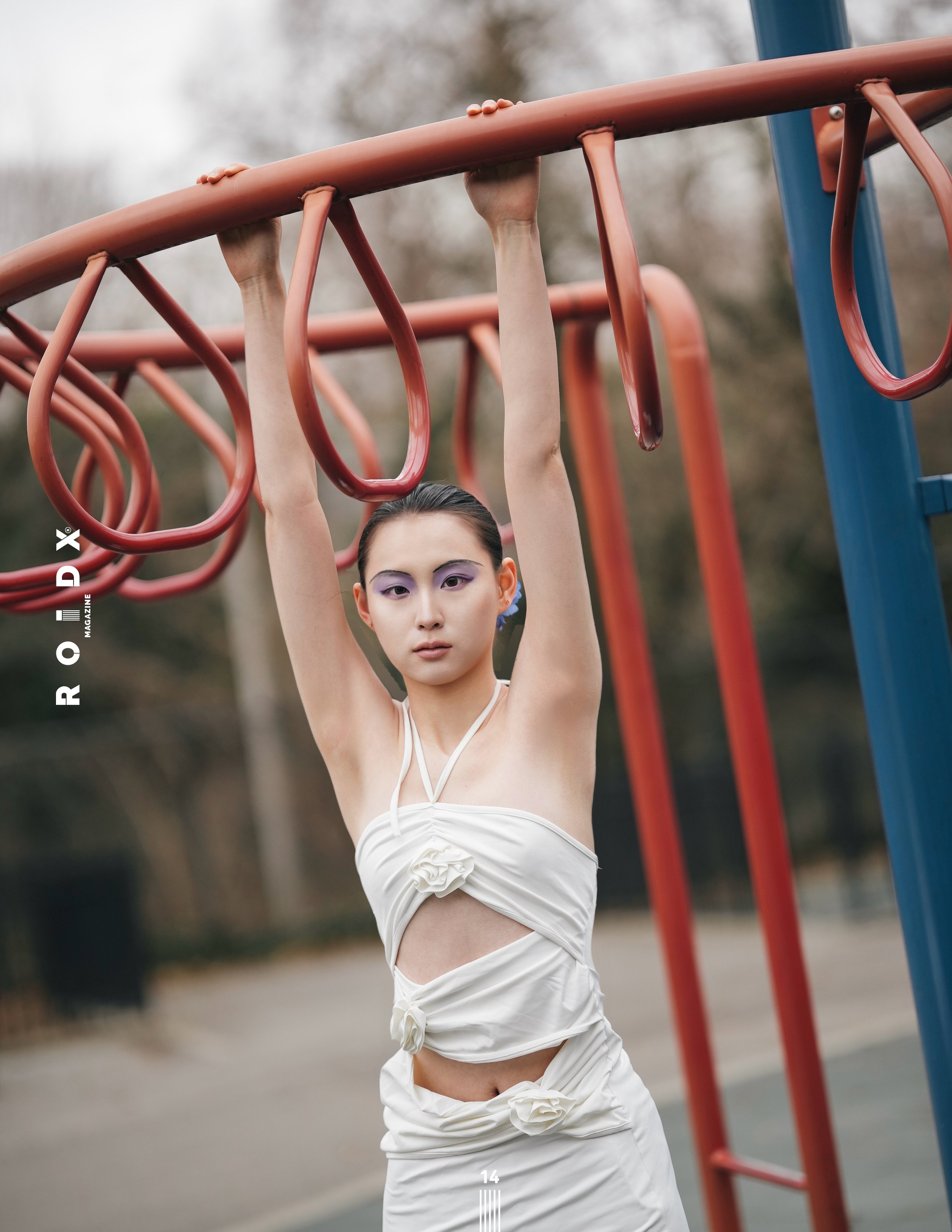 A woman with light skin and dark hair is hanging from a jungle gym in an outdoor park. She is wearing a white dress with a knotted detail at the waist and has purple eyeshadow. The background is blurred trees and sky.