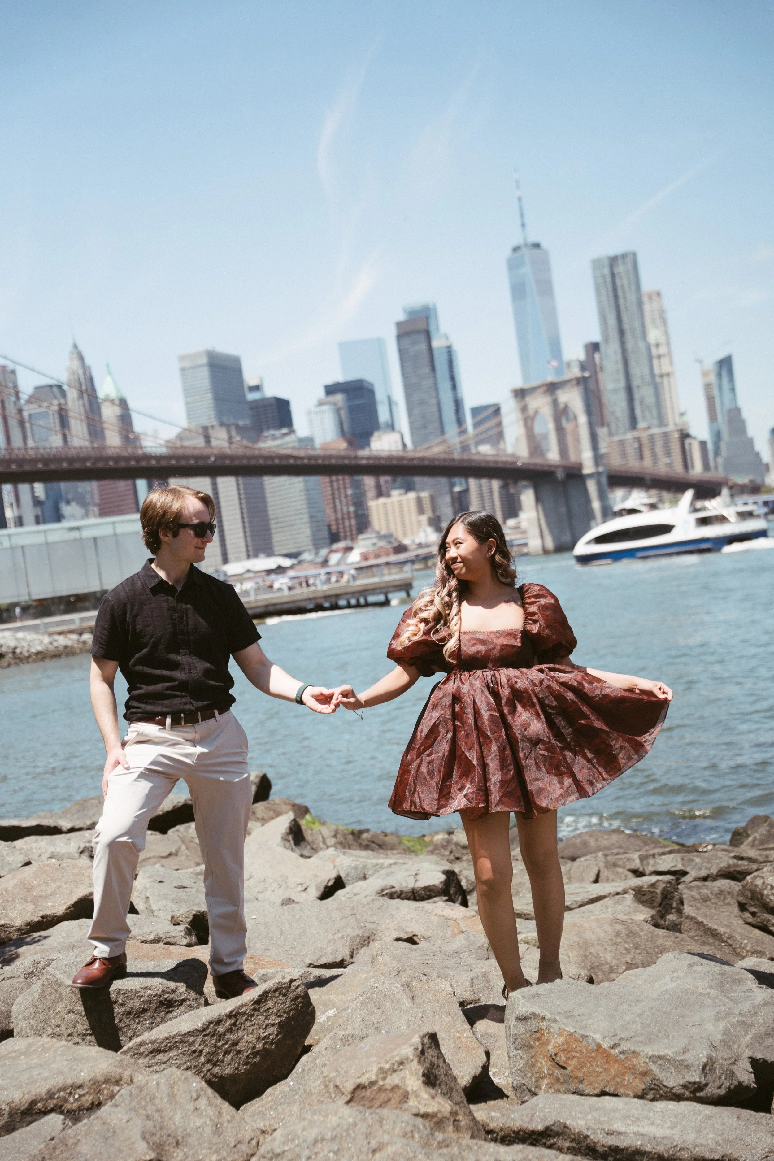 A man and a woman stand on rocks by the water with the New York City skyline, including the Brooklyn Bridge and One World Trade Center, in the background. The man wears sunglasses, a black shirt, and white pants, and the woman wears a vintage-style b