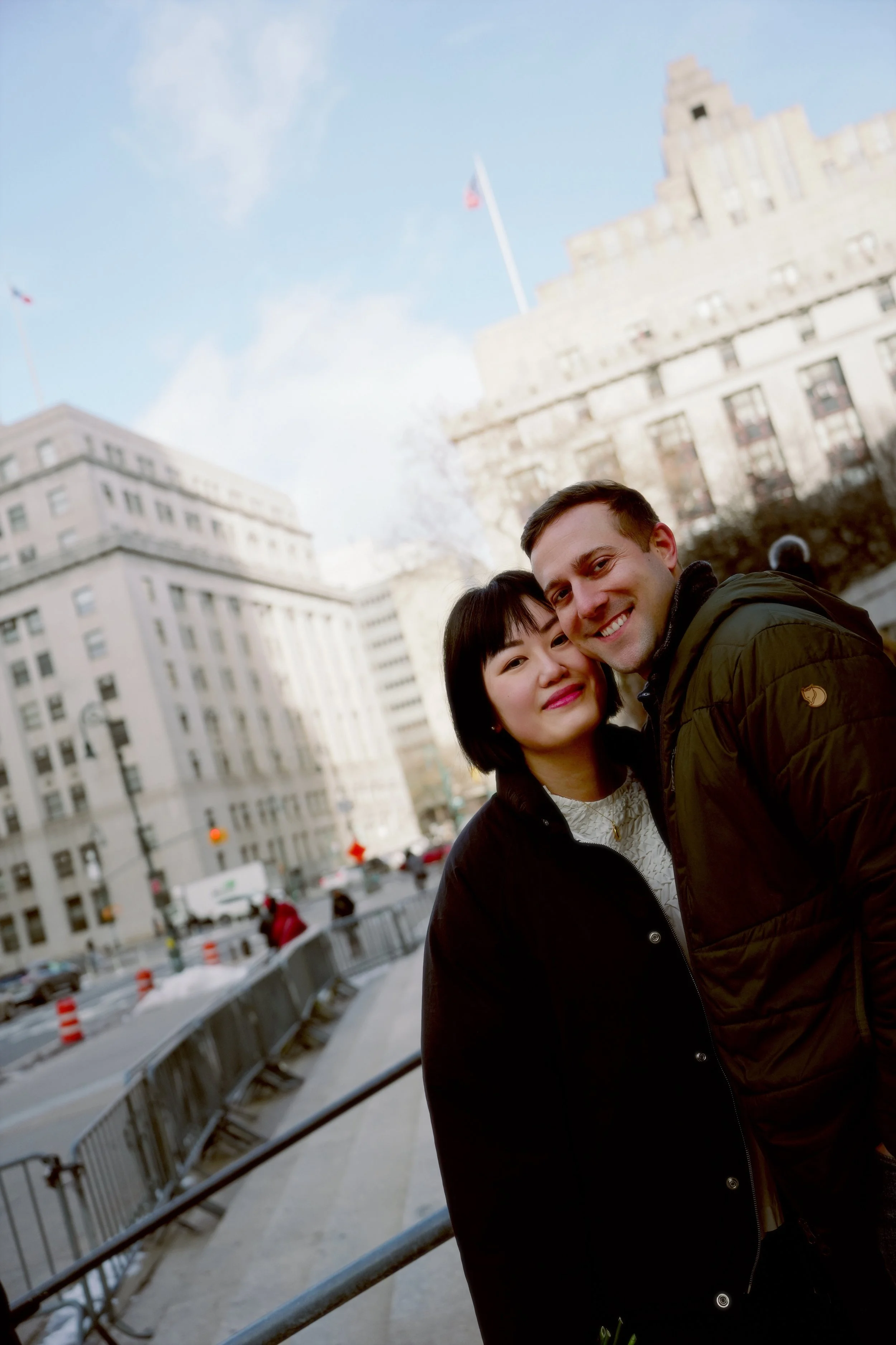 A young man and woman smiling and embracing in an urban setting with big buildings, street barriers, and cars in the background.
