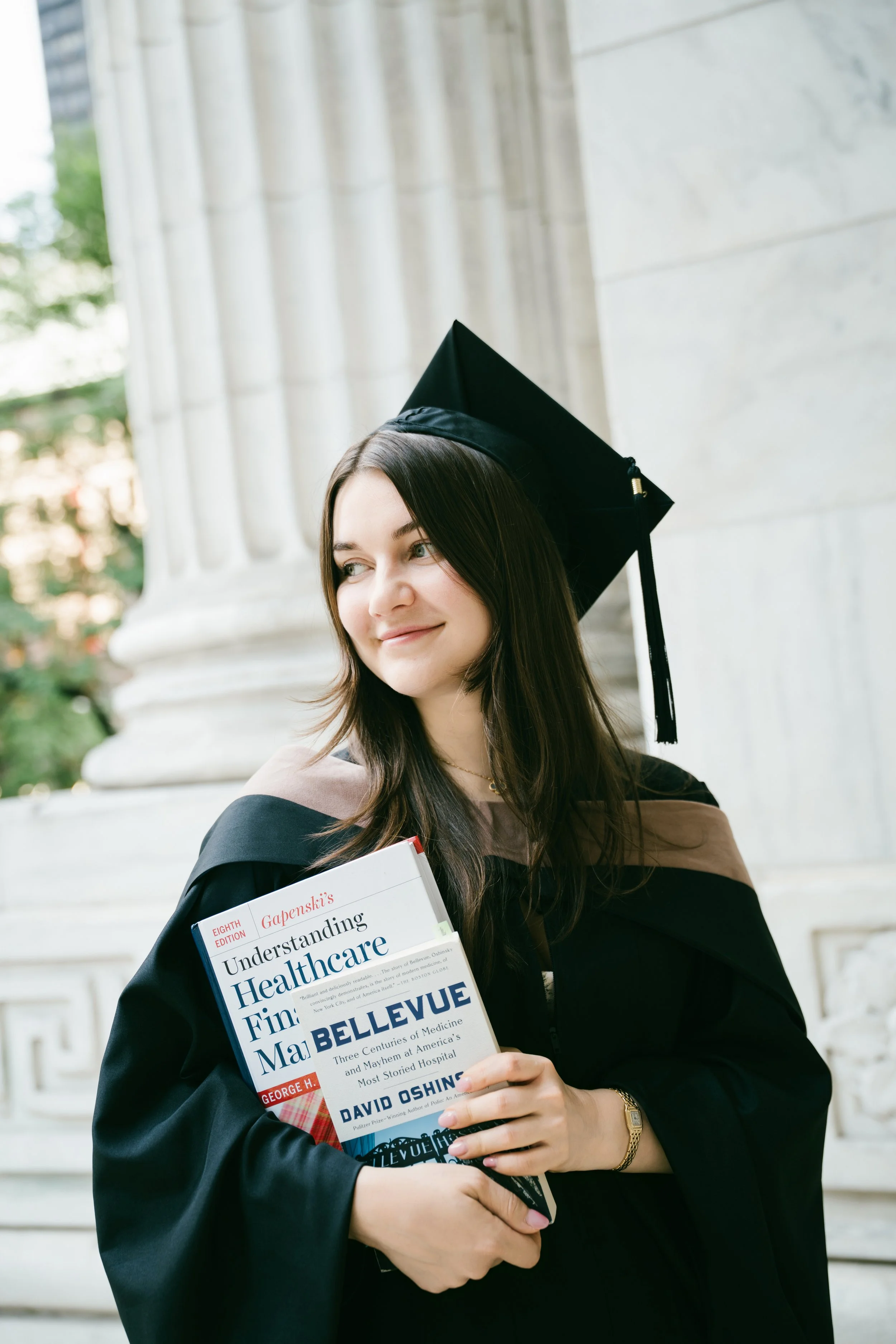 A young woman in a graduation cap and gown holding a book titled 'Understanding Healthcare Finance' outside a building with marble columns.