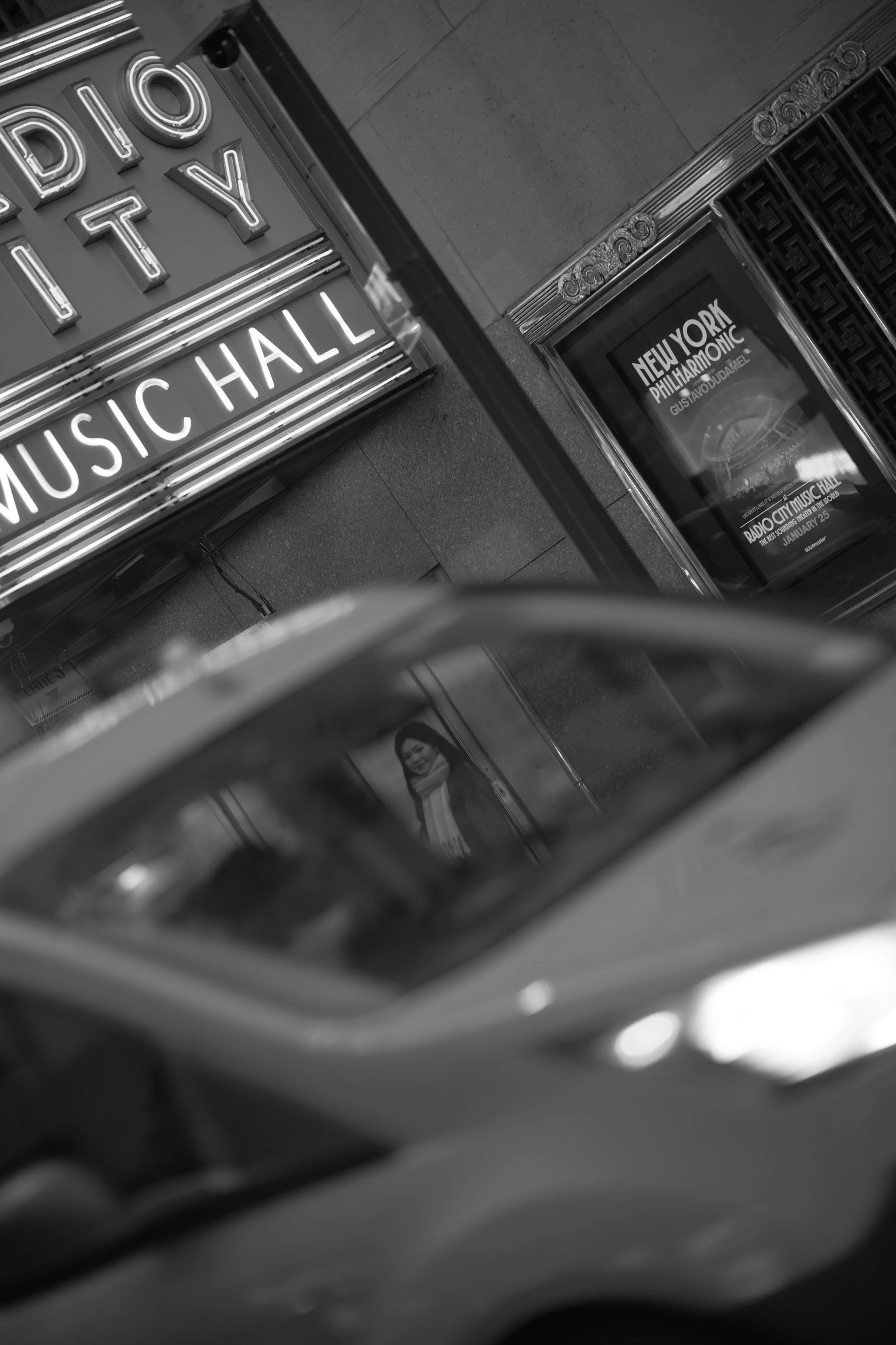 Black and white photo of a subway station entrance with neon sign that reads 'Music Hall' and an advertisement for New York Philharmonic with a photo of a woman's face.