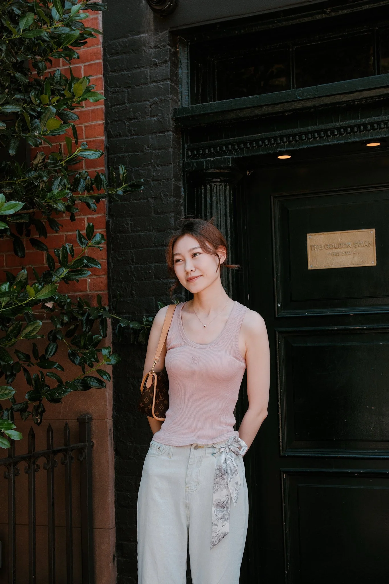 A young woman with short brown hair standing outside near a black door with a gold sign that reads 'The Golden Swan EST 2023'. She is wearing a sleeveless beige top, white pants, and carrying a Louis Vuitton bag. There are green leaves on her left si