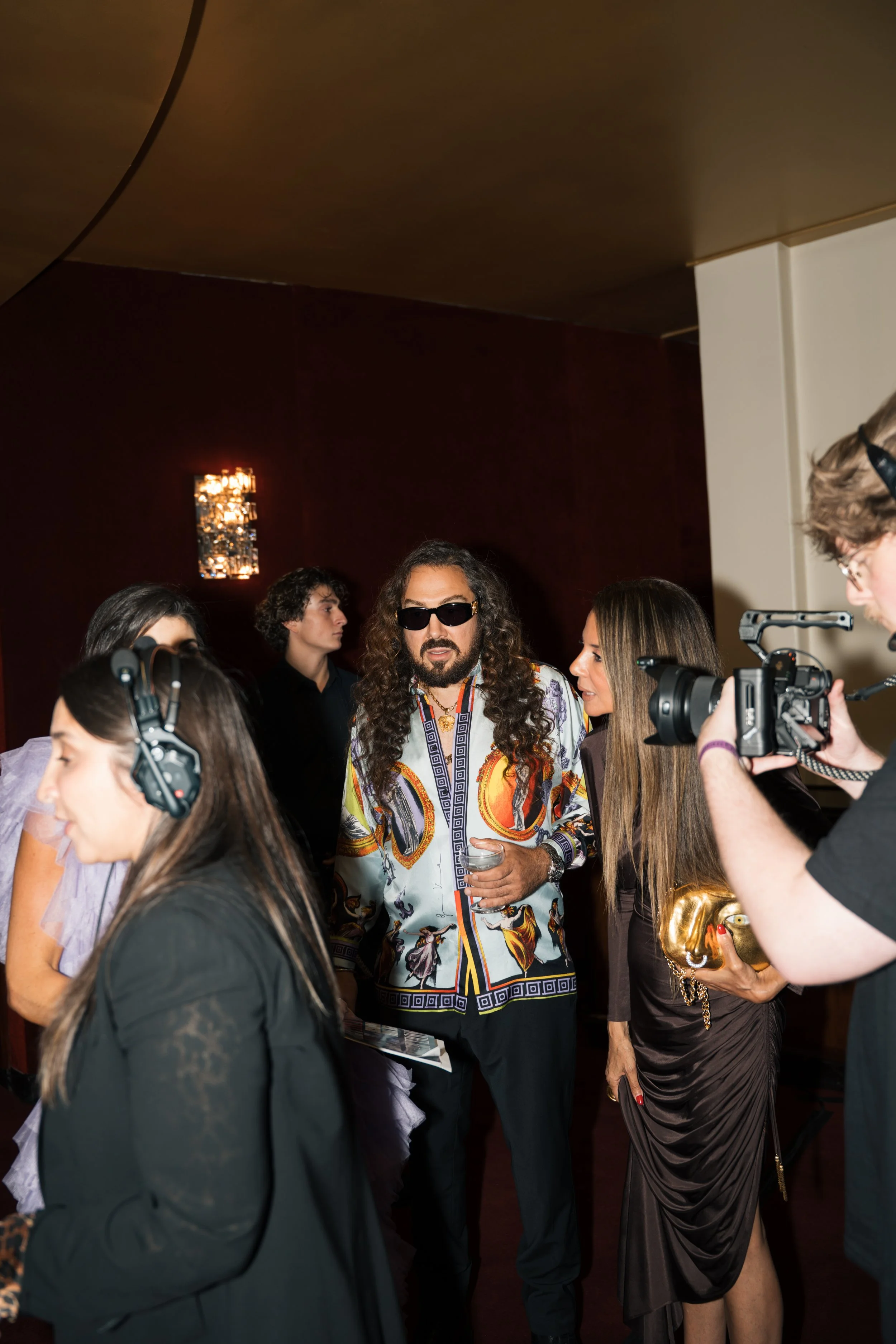 Group of people at an indoor event, some wearing headphones, and one man with long curly hair and sunglasses in the center.