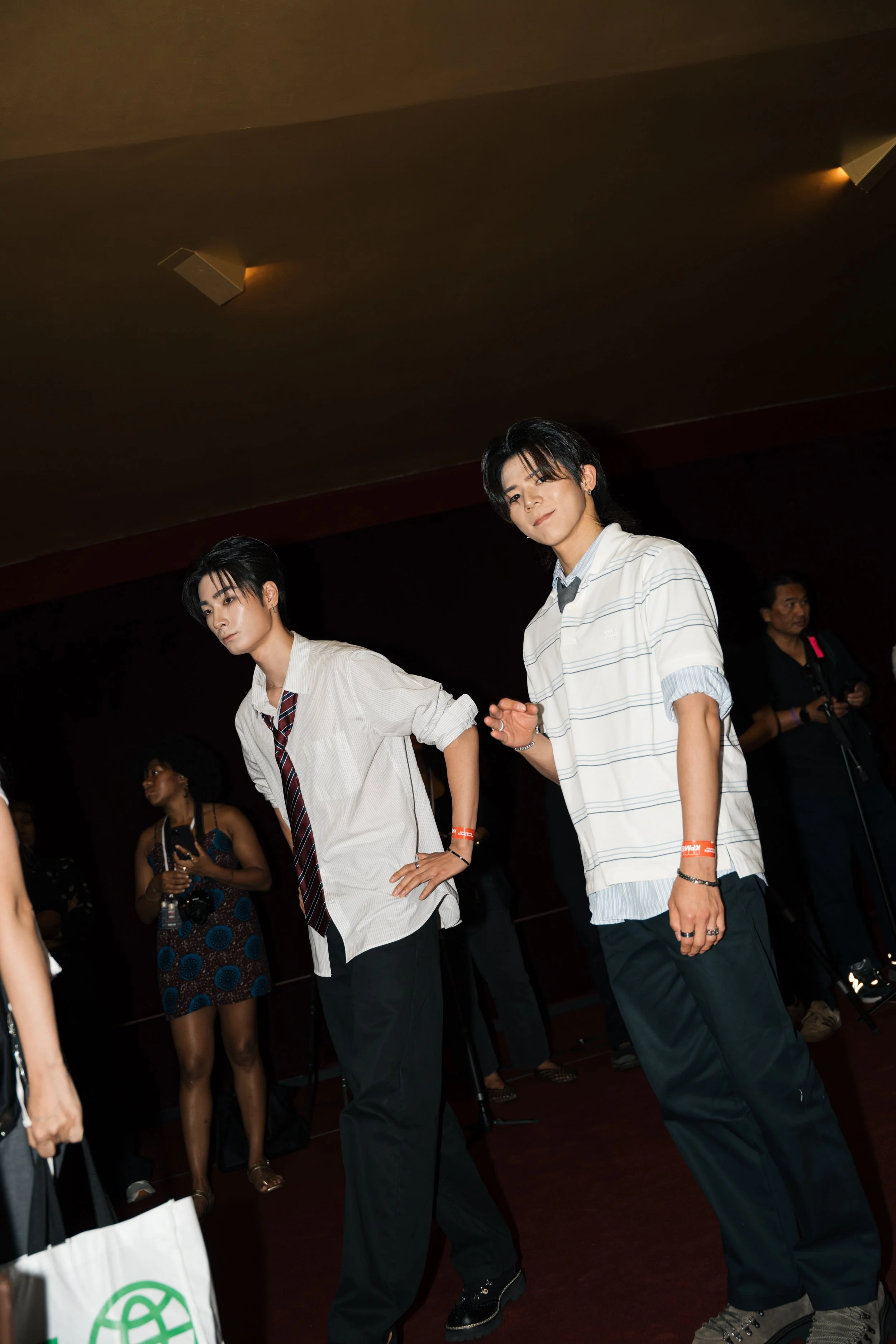 Two young men with dark hair wearing casual school uniforms, walking and talking in a darkened indoor space. One is wearing a white shirt with black pants and a tie, the other a striped shirt with jeans.