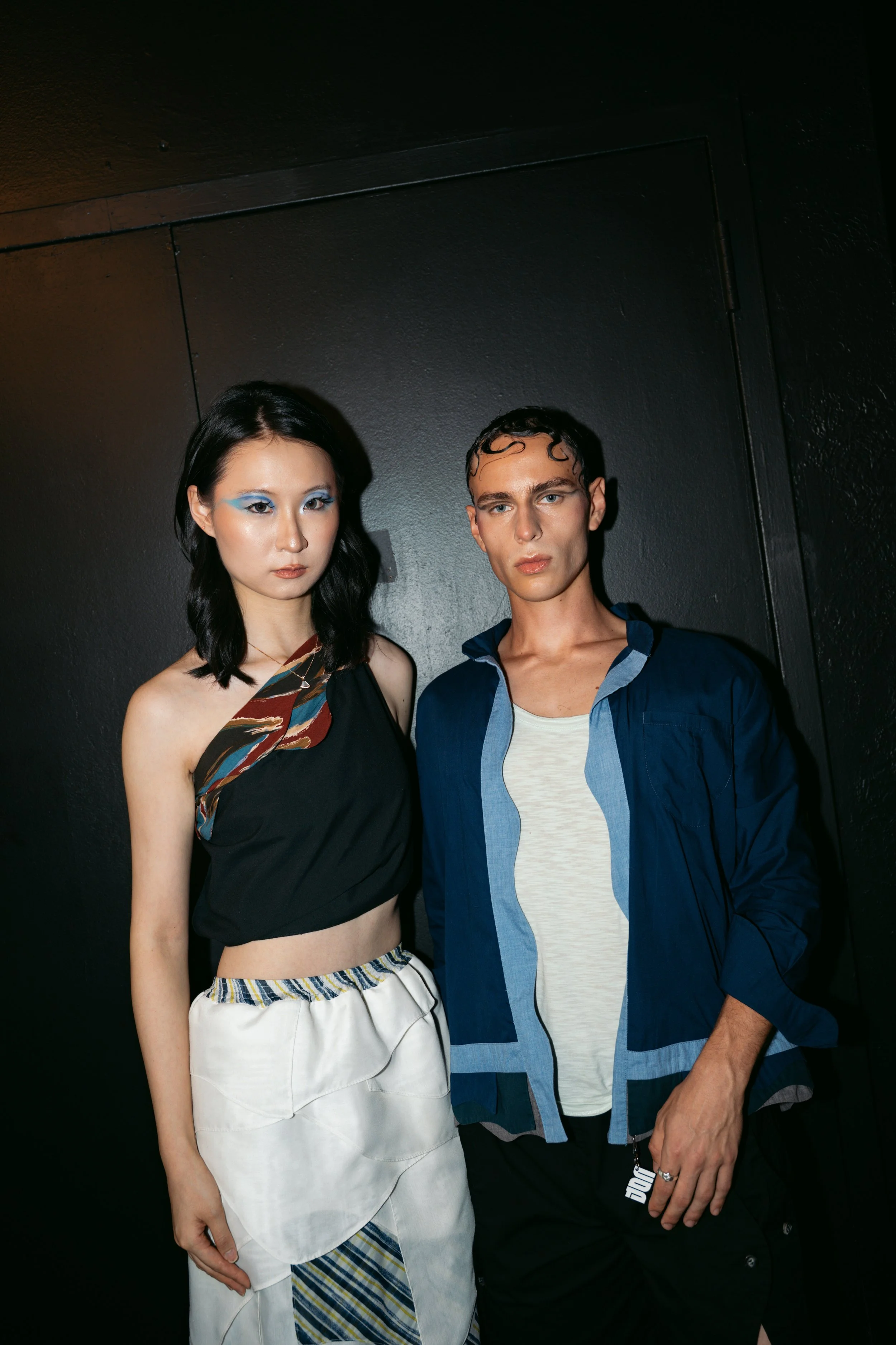 Two young adults, a woman with black hair and a man with wet, curly hair, stand together against a black wall. The woman wears a stylish black top with colorful patterns and white wide-leg pants with cloudy patterns. The man is dressed in a blue jack