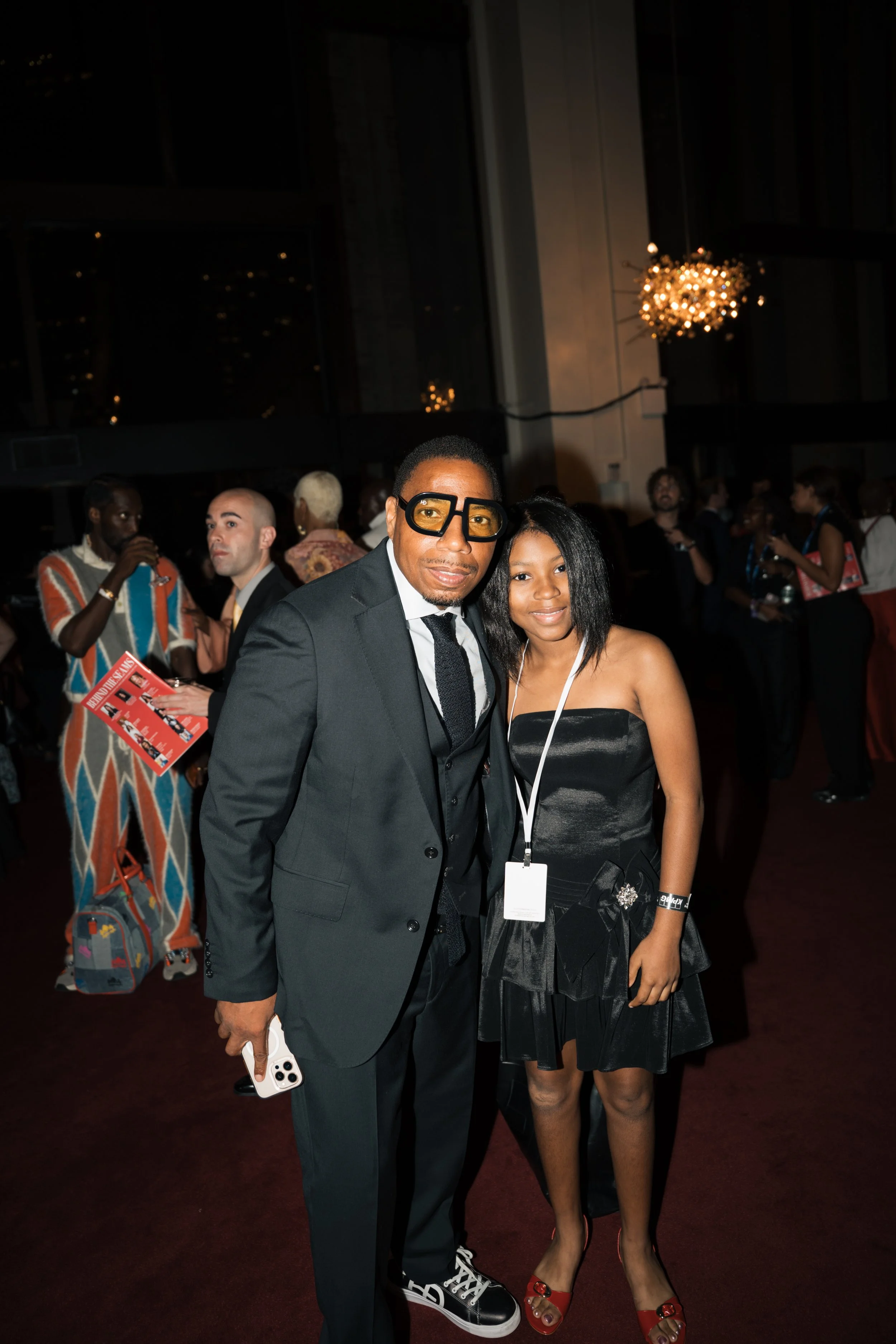 Man in black suit and yellow glasses standing next to young woman in black dress and red sandals at an indoor event, with other guests in the background.