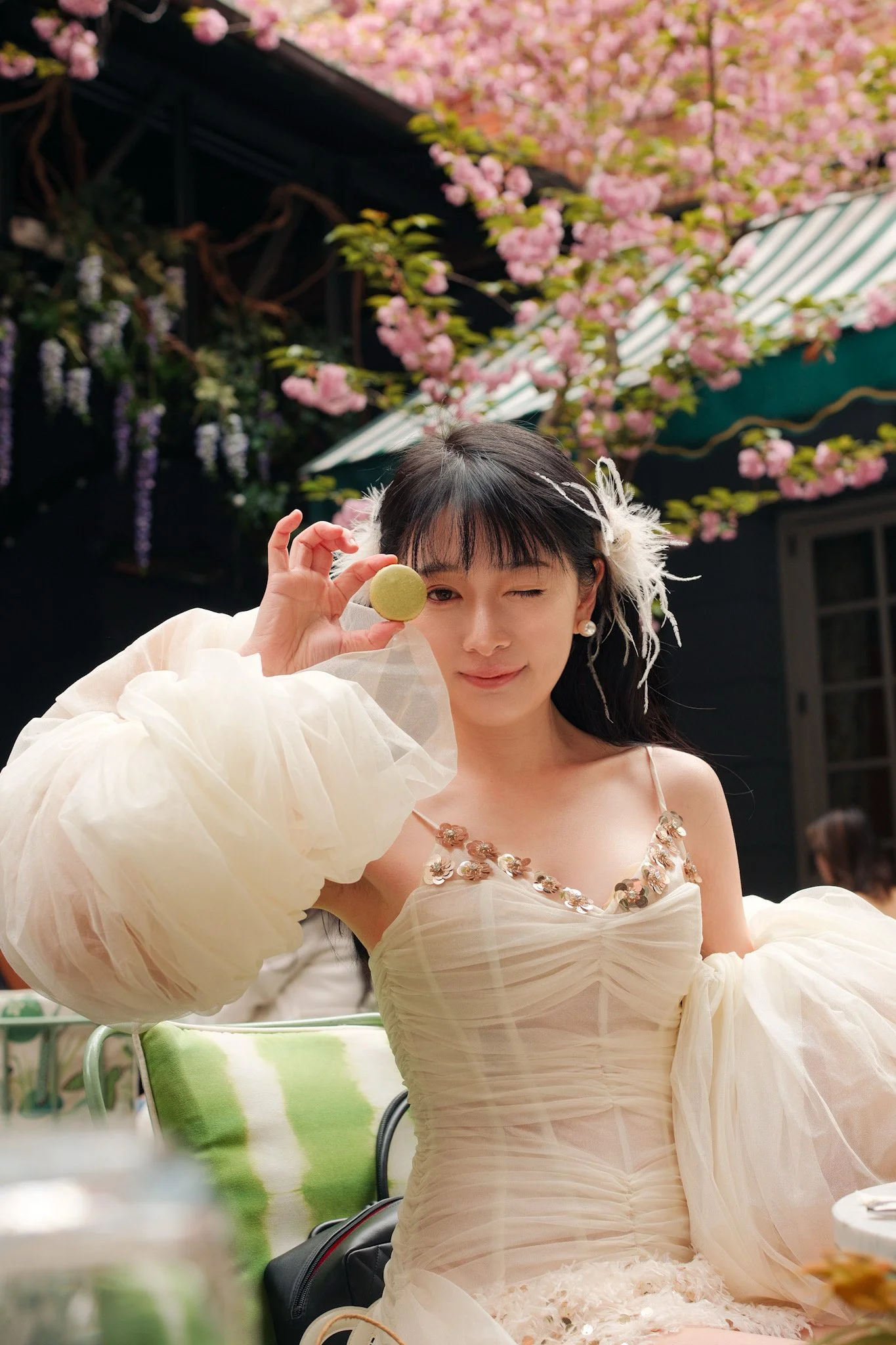 A woman dressed in a light beige gown with decorative embellishments on the bodice, holding a green macaron near her eye and winking. She has dark hair with a feathered hair accessory and pearl earrings, standing outdoors with pink flowering trees, a