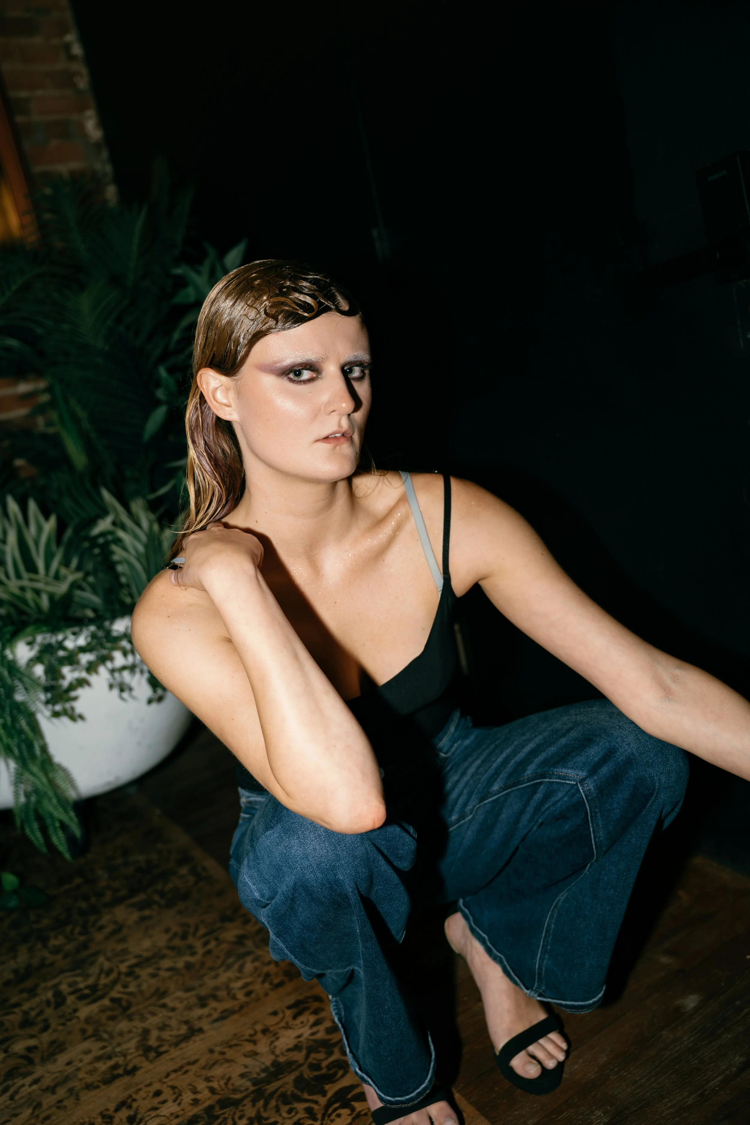 A woman with wet hair, wearing a black spaghetti strap top and blue jeans, sitting on the floor with one knee up and the other leg bent. She has a serious expression and is touching her neck with her right hand. There are green plants behind her, and