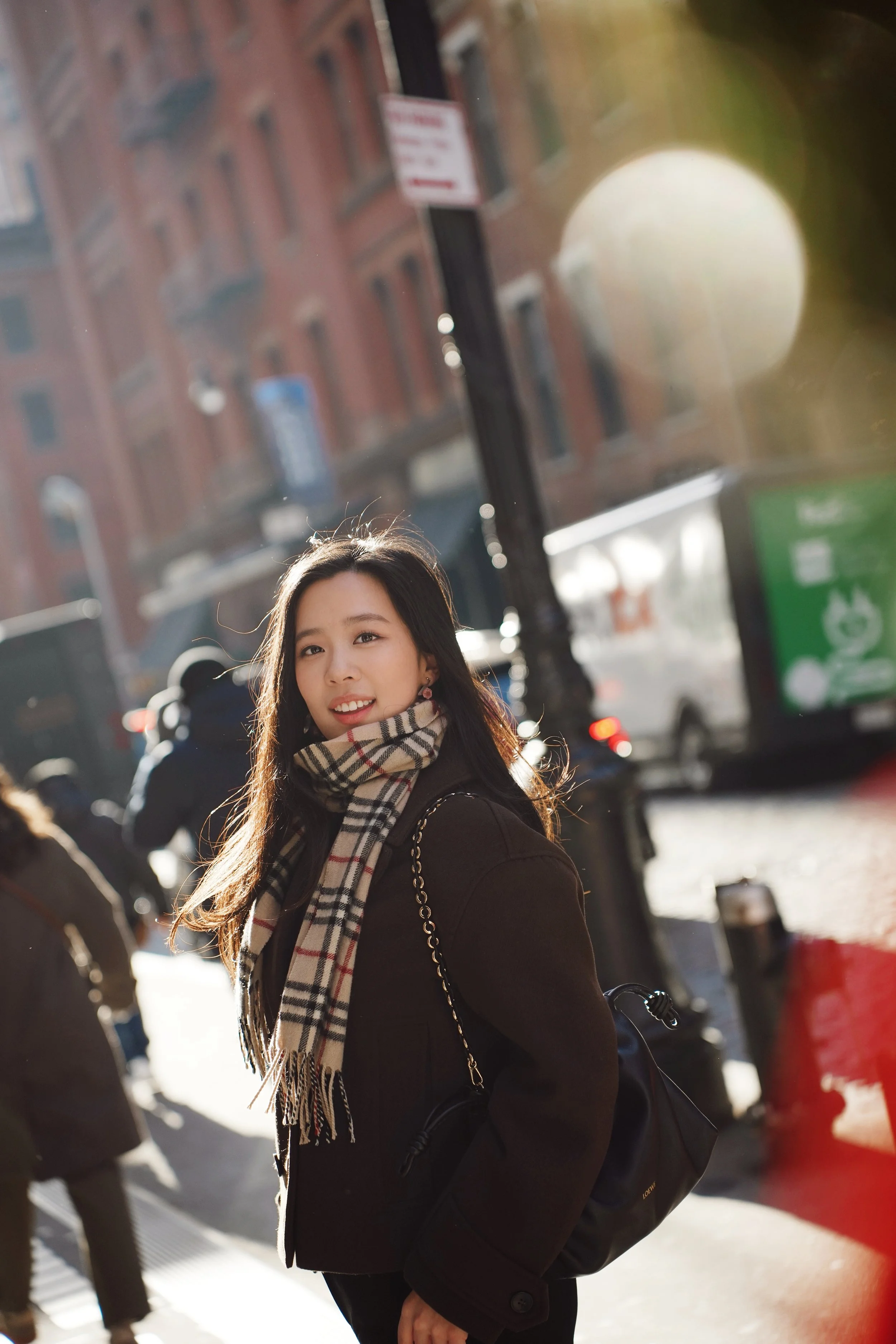 A woman with long dark hair, smiling, wearing a black coat, patterned scarf, and carrying a black handbag, standing on a busy city street with buildings and pedestrians in the background.