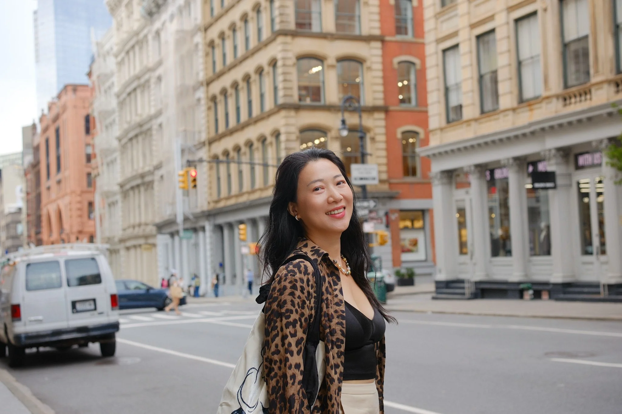 A woman with long dark hair smiling in an urban street with historic brick and stone buildings, cars, and a traffic light in the background.
