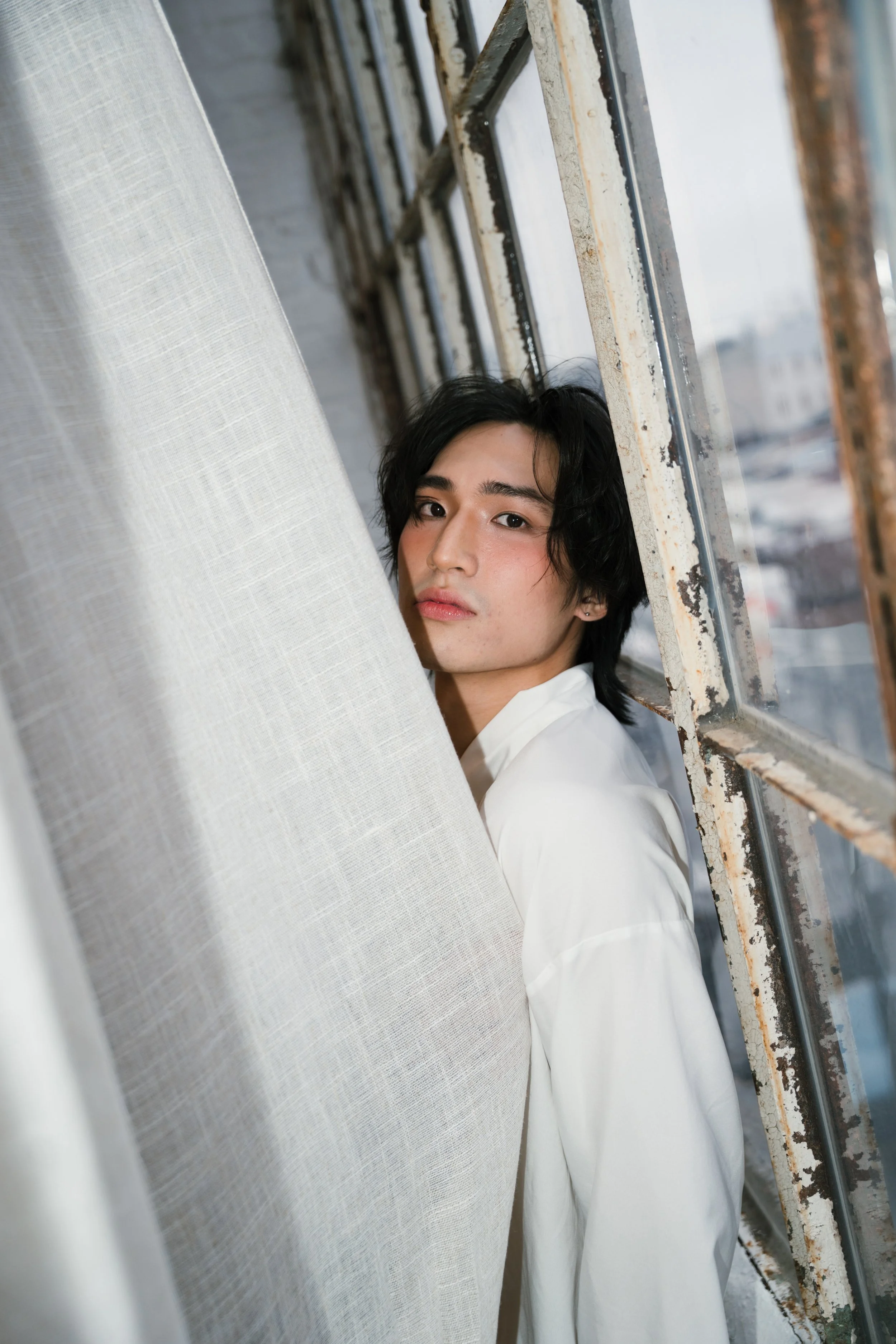 Young person with dark hair looking out a large window with a rusted metal frame, partially obscured by a white curtain, in an industrial-style setting.