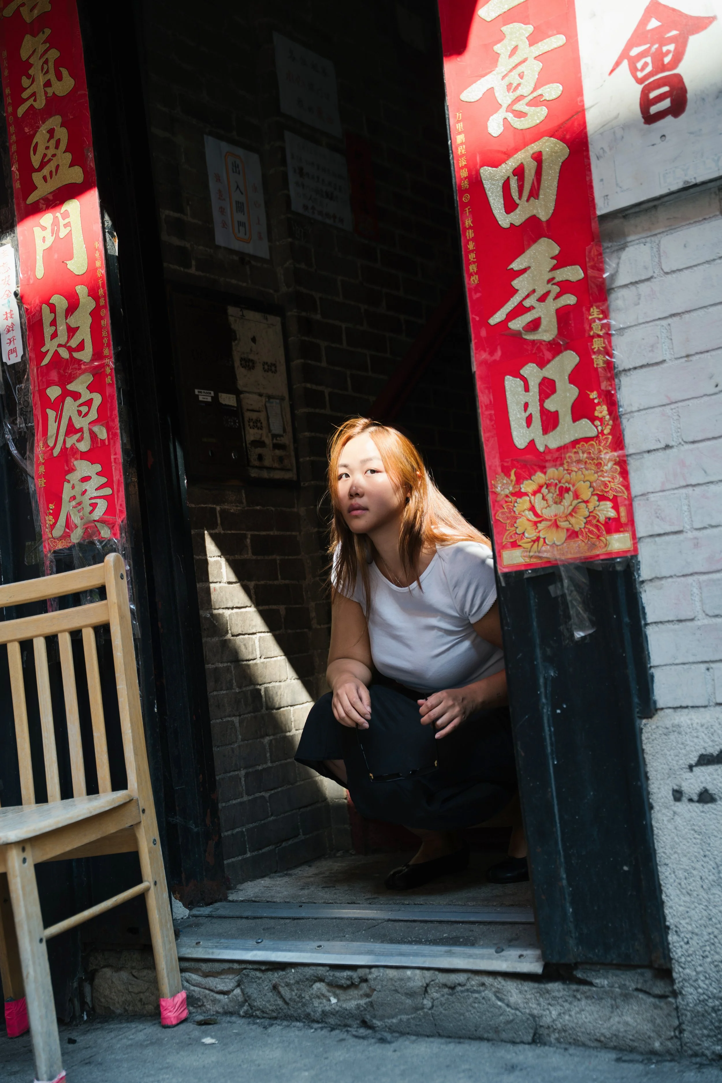 A young woman with light brown hair crouching in the doorway of a building with red and gold Chinese banners hanging beside the entrance.
