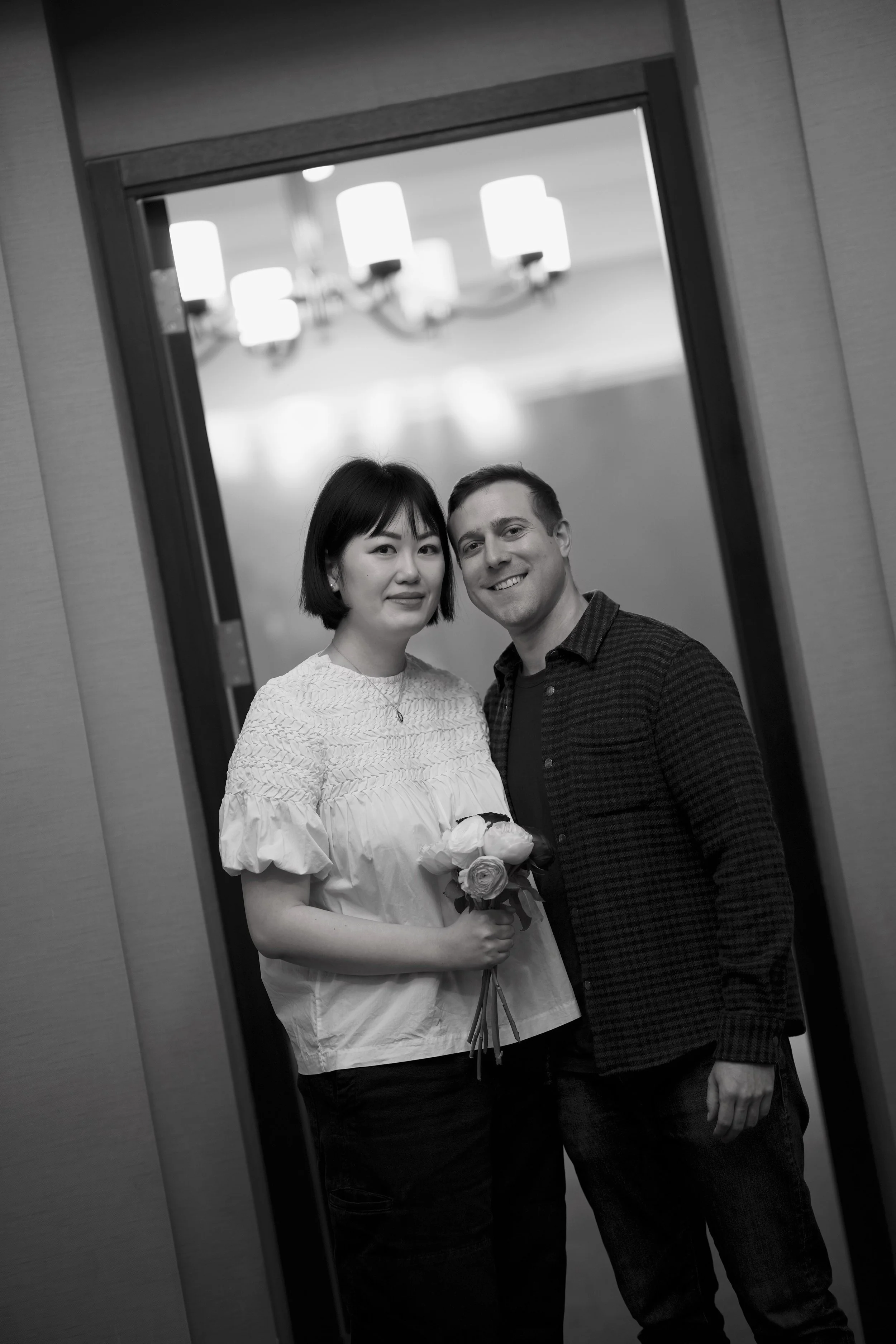 A black-and-white photo of a couple reflected in a mirror. The woman is holding a bouquet of roses and is wearing a light-colored top. The man is dressed in a striped shirt and has his arm around her. They appear to be at a formal event or celebratio