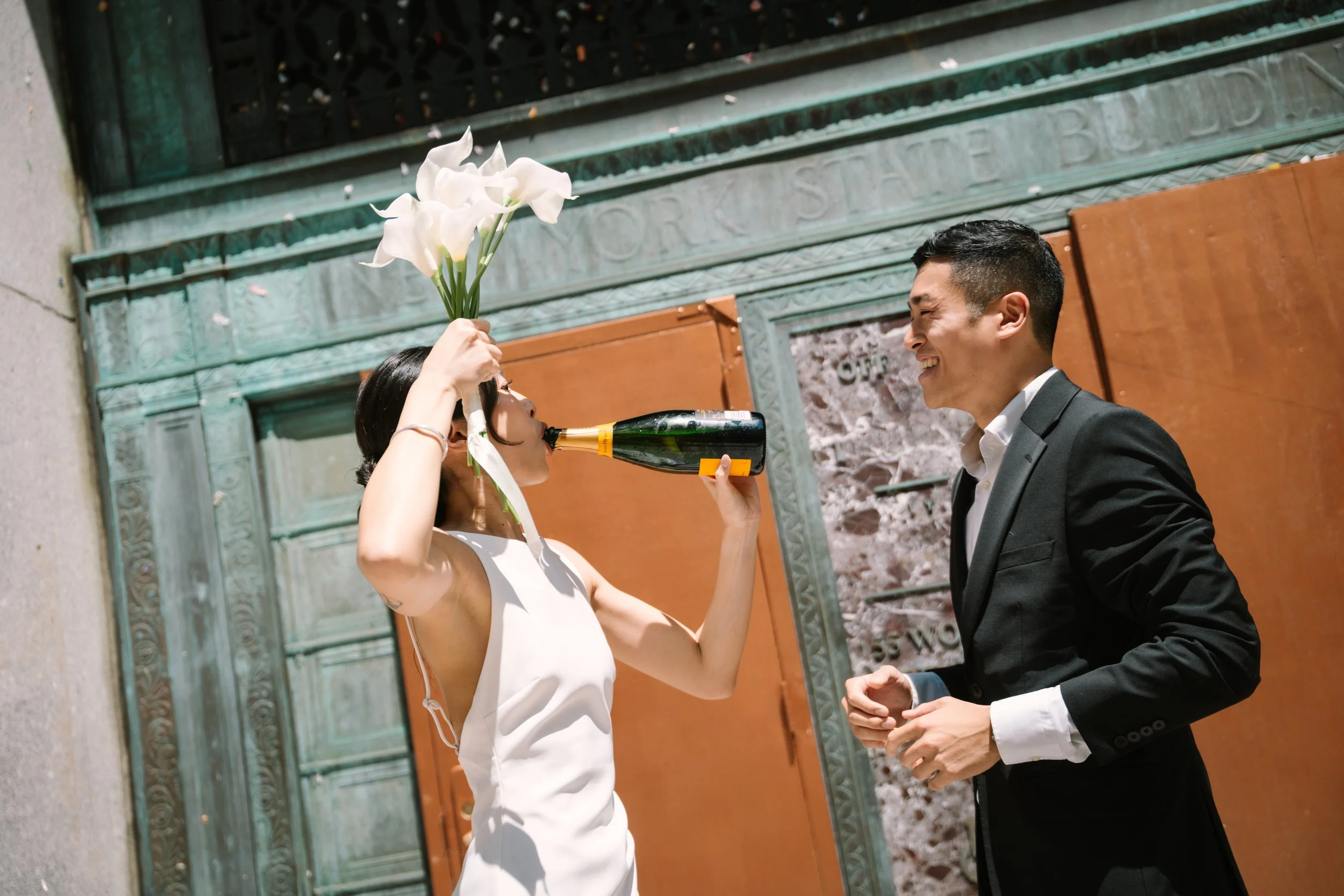 A woman in a white dress holding a bouquet of white calla lilies over the bottle of champagne aimed at a smiling man in a tuxedo, outside in front of a green ornate building with a wooden door.