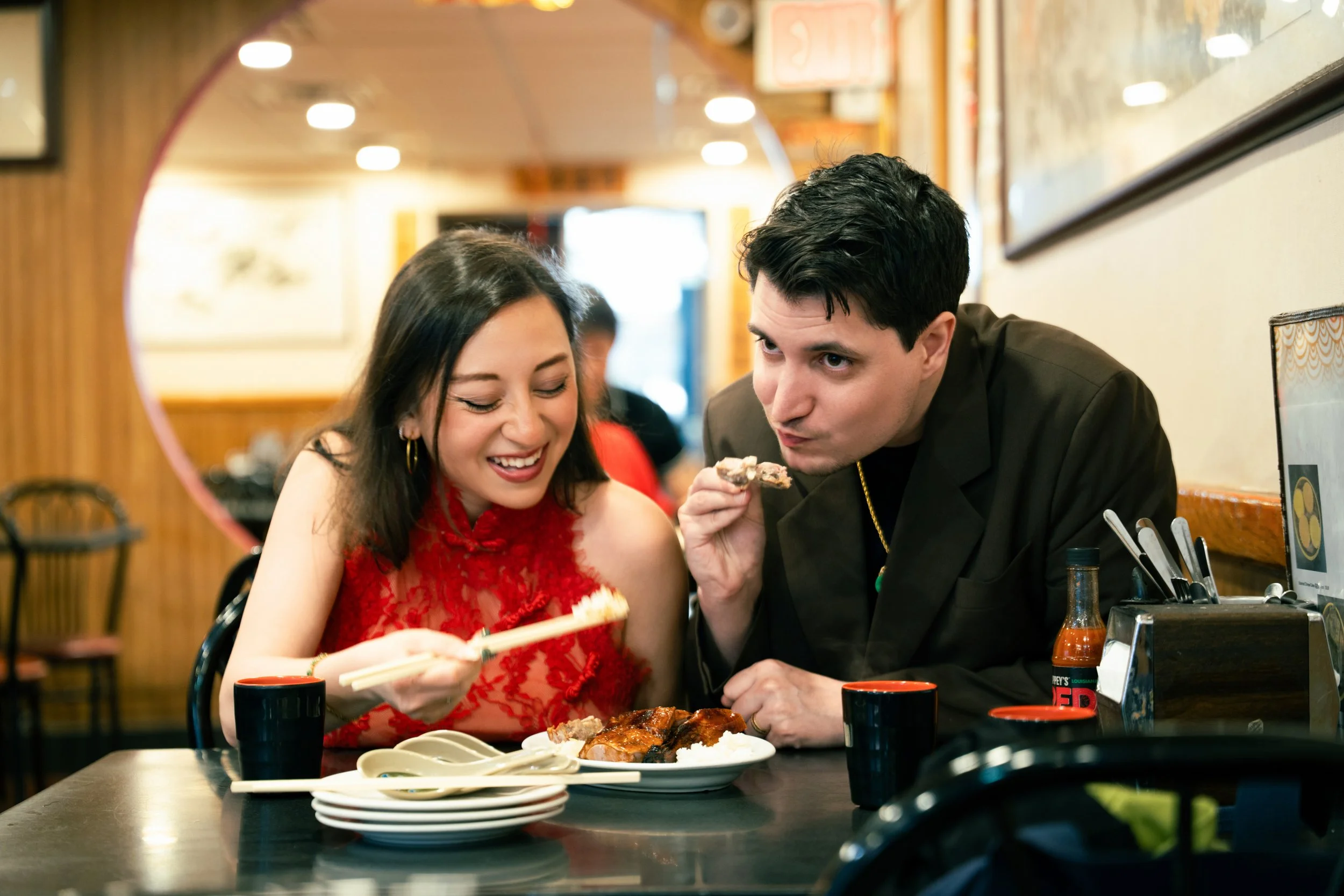A young woman in a red dress and a young man in a dark jacket sharing a meal at a restaurant, smiling and enjoying each other's company.