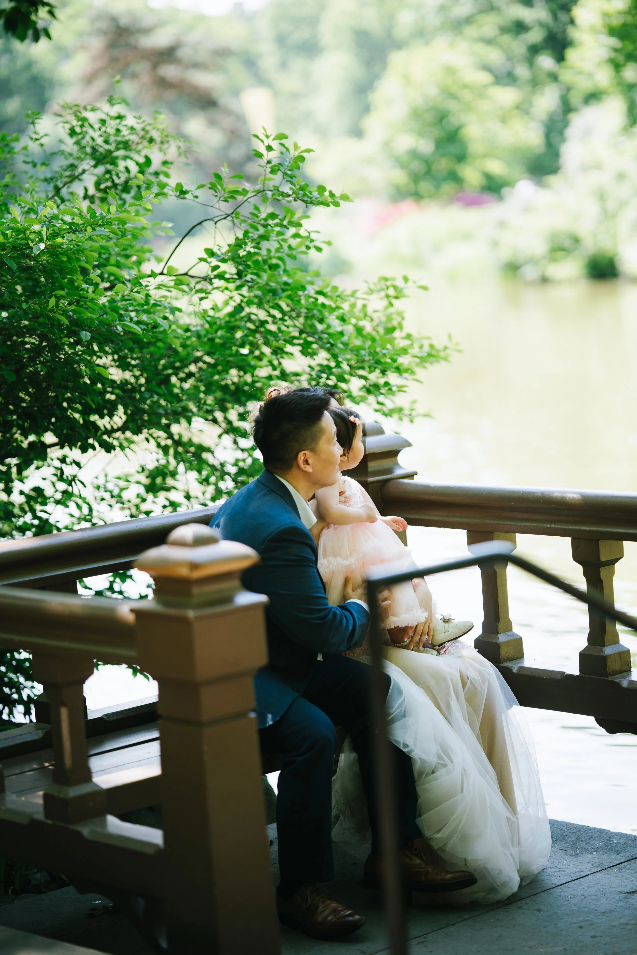 A man in a blue suit and a young girl in a wedding dress are sitting on a park bench by a body of water, surrounded by lush green trees.
