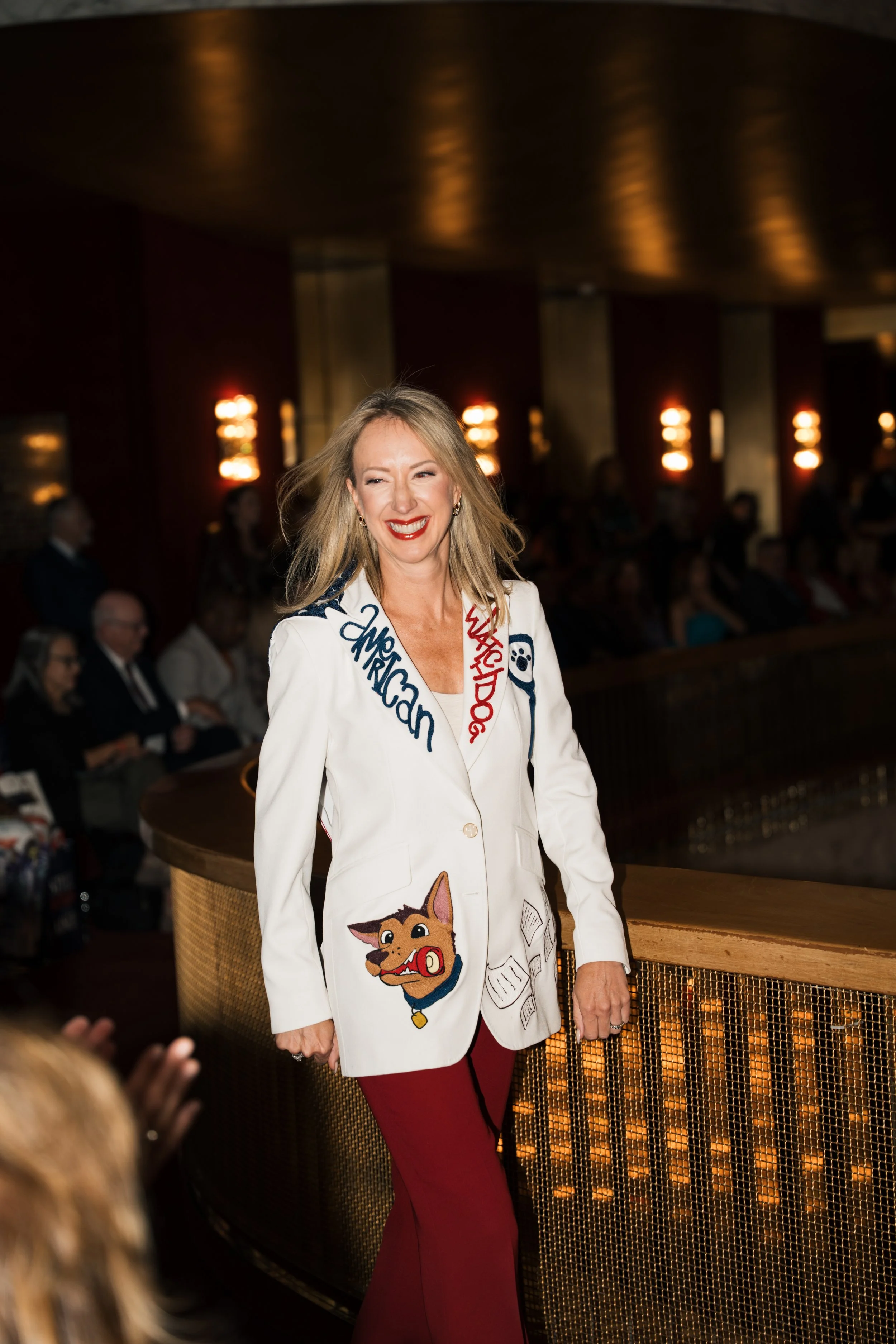 A woman with blonde hair, red lipstick, and a Aries-themed blazer smiling and walking in a dimly lit room with audience members in the background.