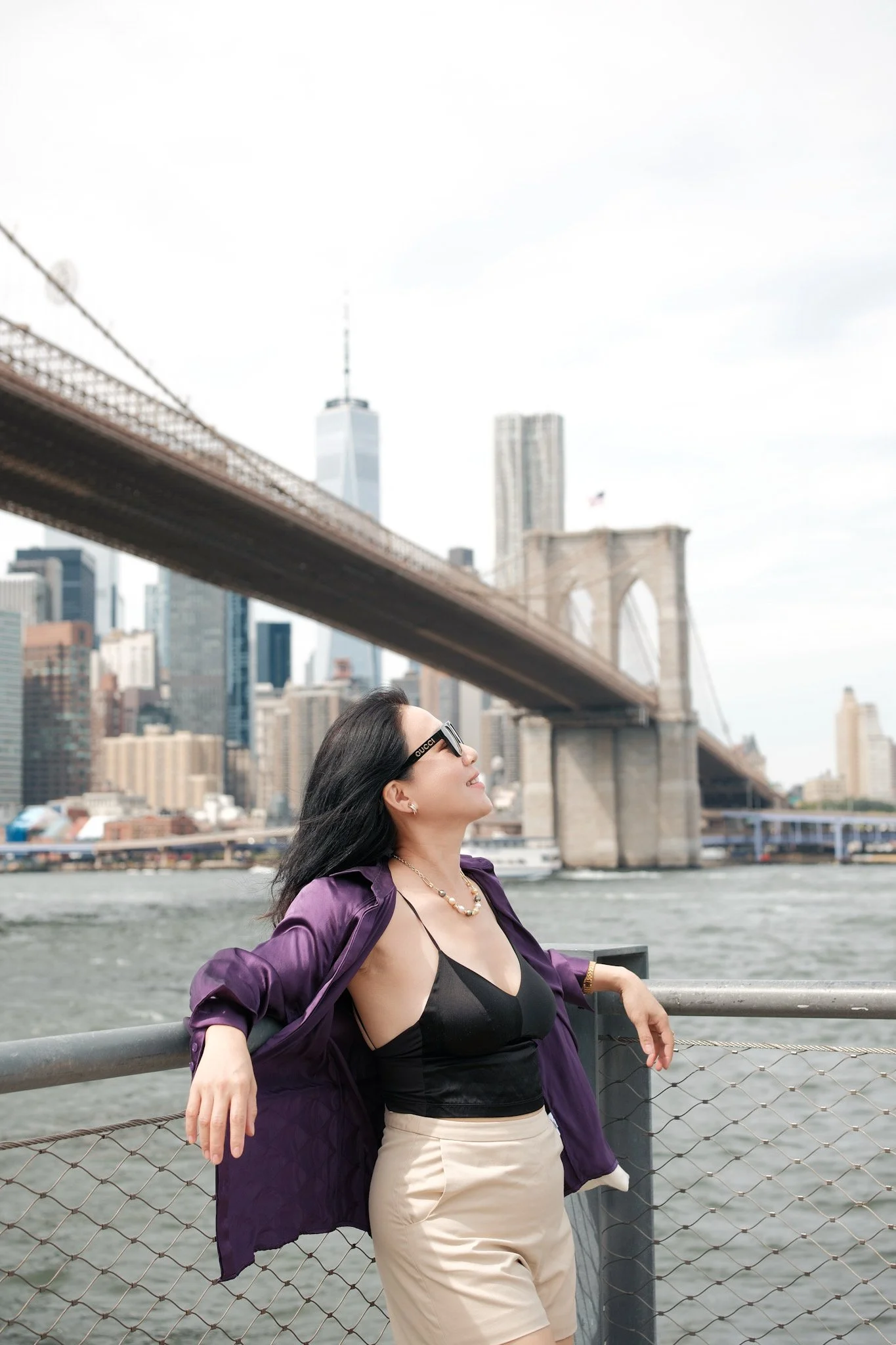 A woman with long dark hair wearing sunglasses, a black top, beige high-waisted pants, and a purple jacket draped over her shoulders, leaning on a railing by the water with the Brooklyn Bridge and city skyline in the background.